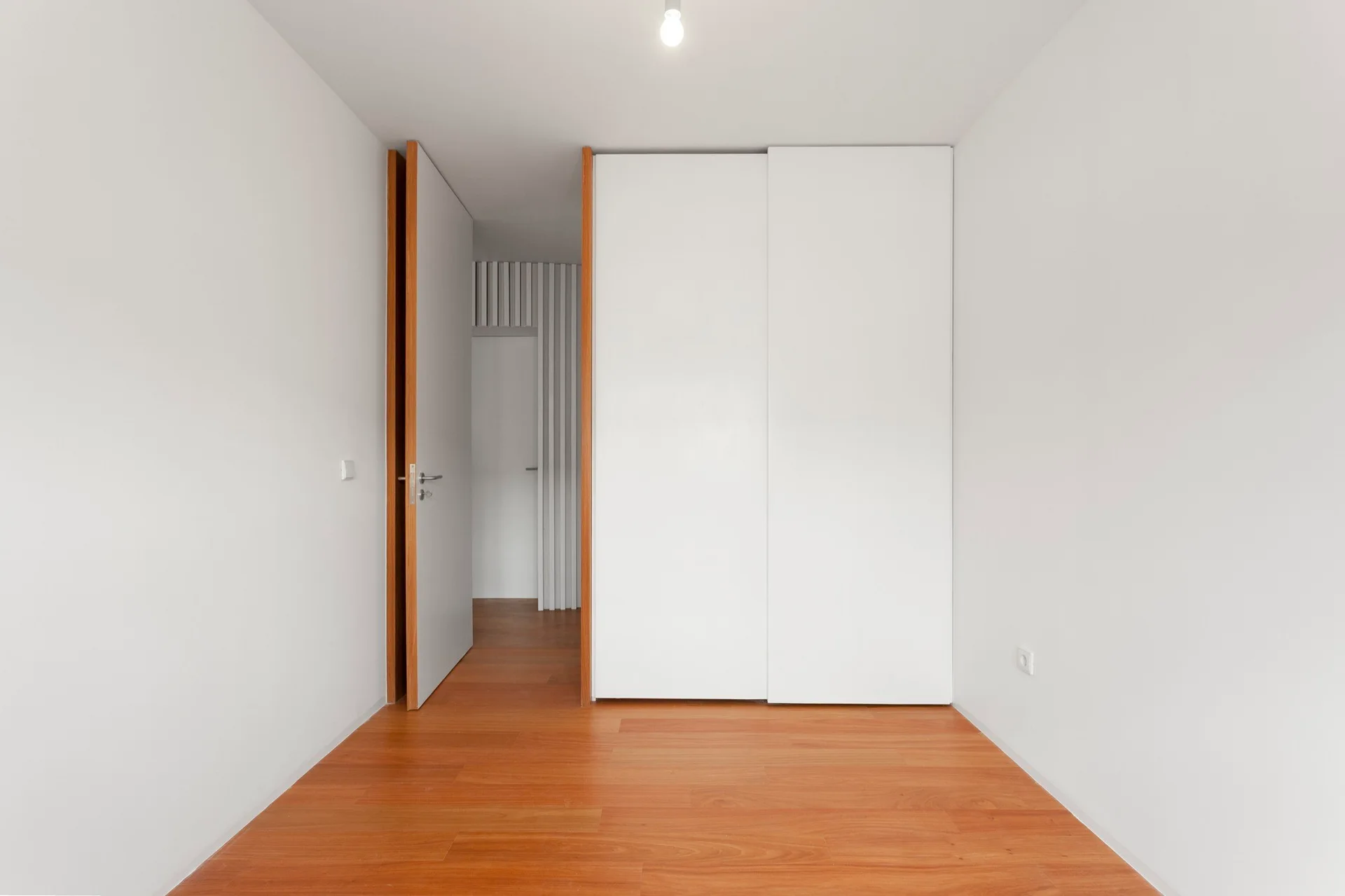 A minimalist bedroom interior features warm wood plank flooring, a white built-in wardrobe with sliding doors, and an open entryway.