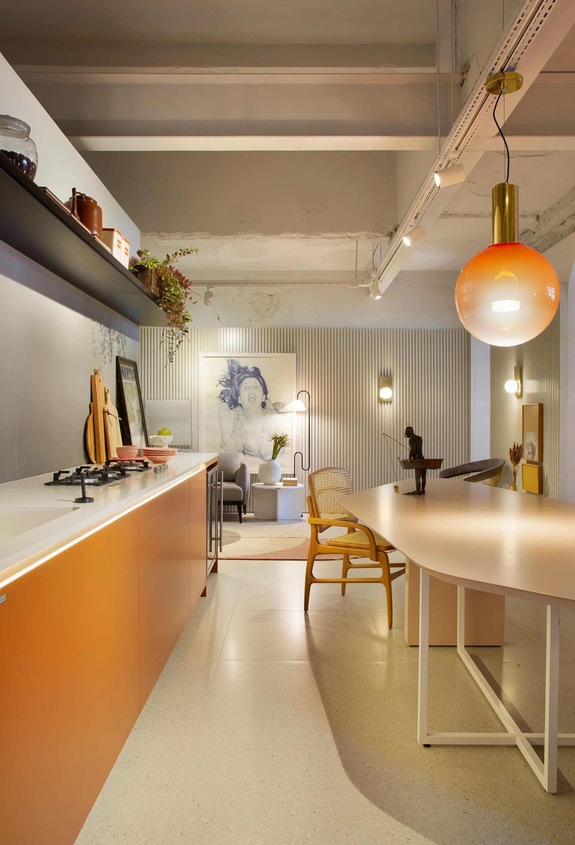 An open kitchen with matte orange cabinets, a light dining table with cane-back chairs, and a white vertically ribbed accent wall.