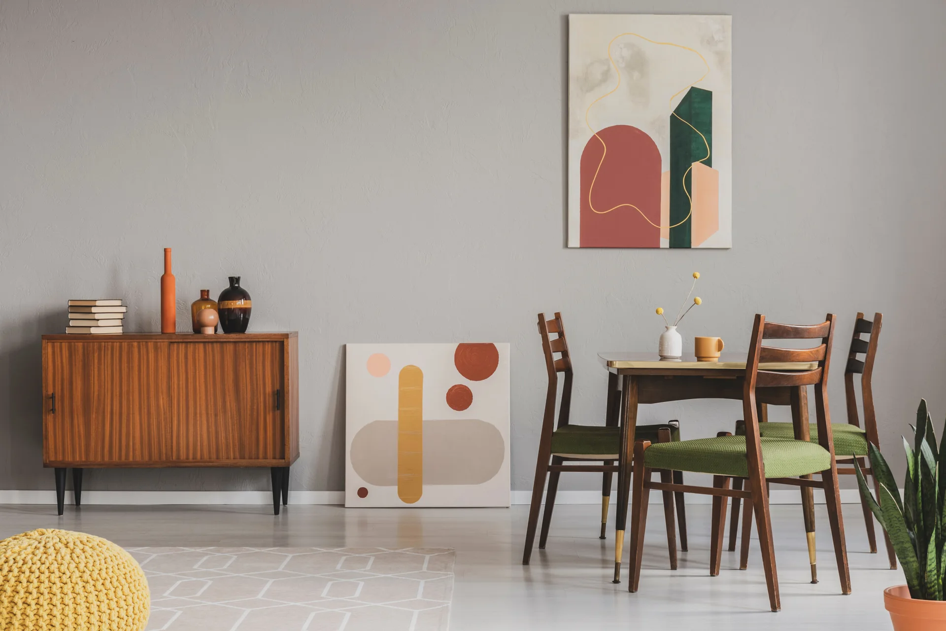 A mid-century modern dining room features a wooden dining set with green chairs, a vintage sideboard, and abstract paintings against a grey wall.