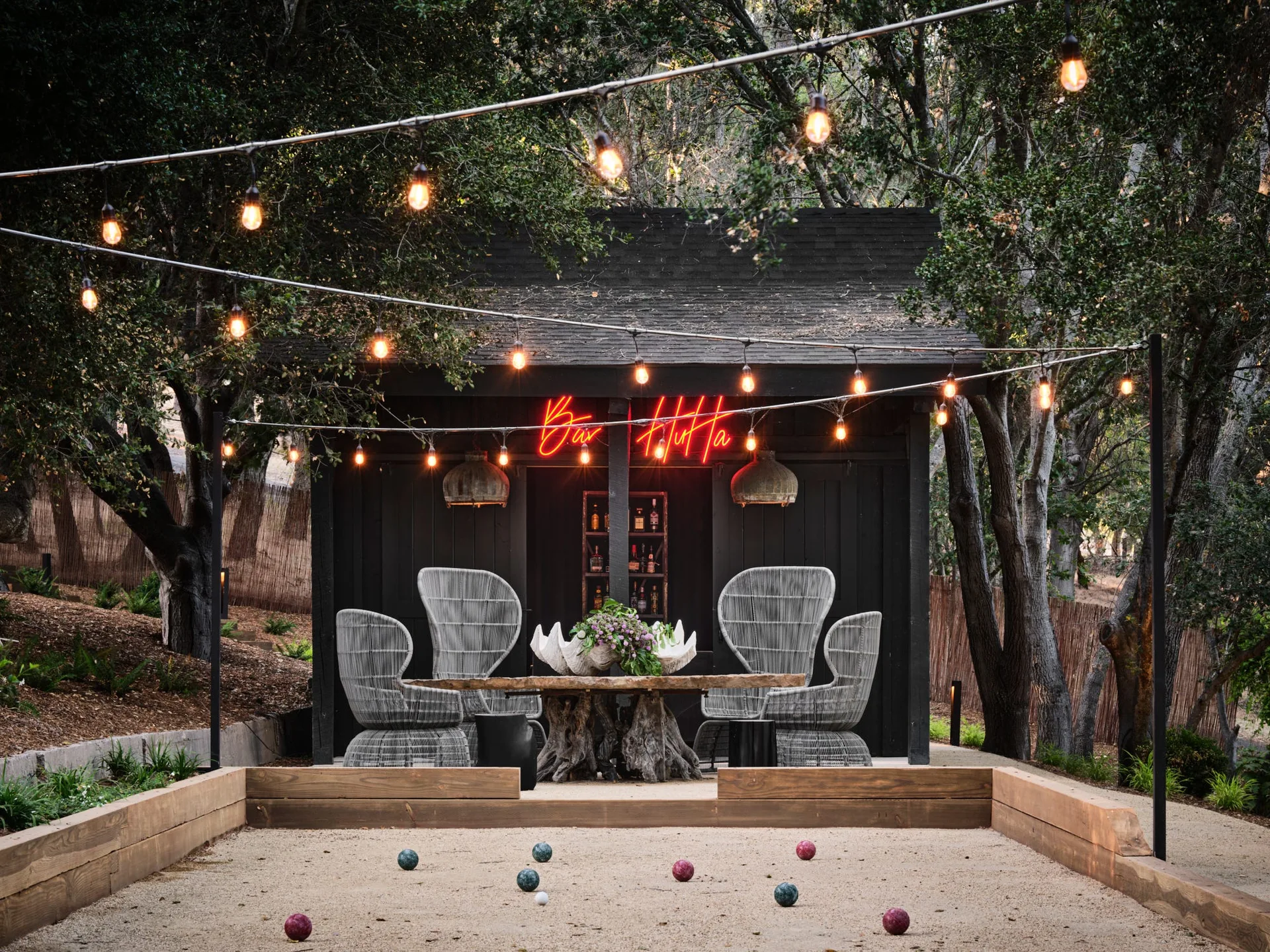 An outdoor bocce ball court sits in front of a black wooden bar shed featuring woven armchairs, a live-edge dining table, and glowing overhead string lights.