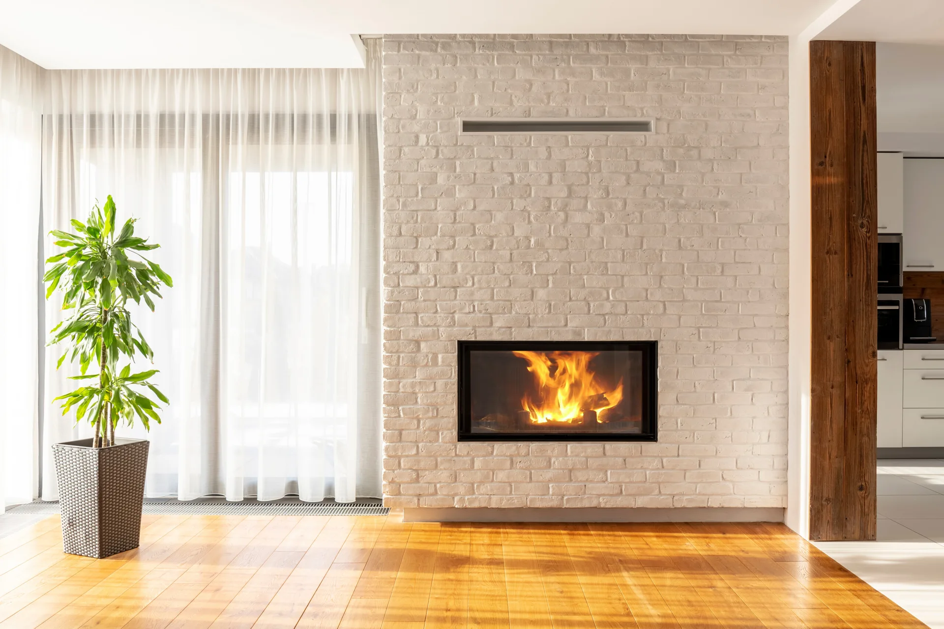A modern living space features a burning fire in a white brick fireplace, warm hardwood floors, a green houseplant, and sheer white curtains.