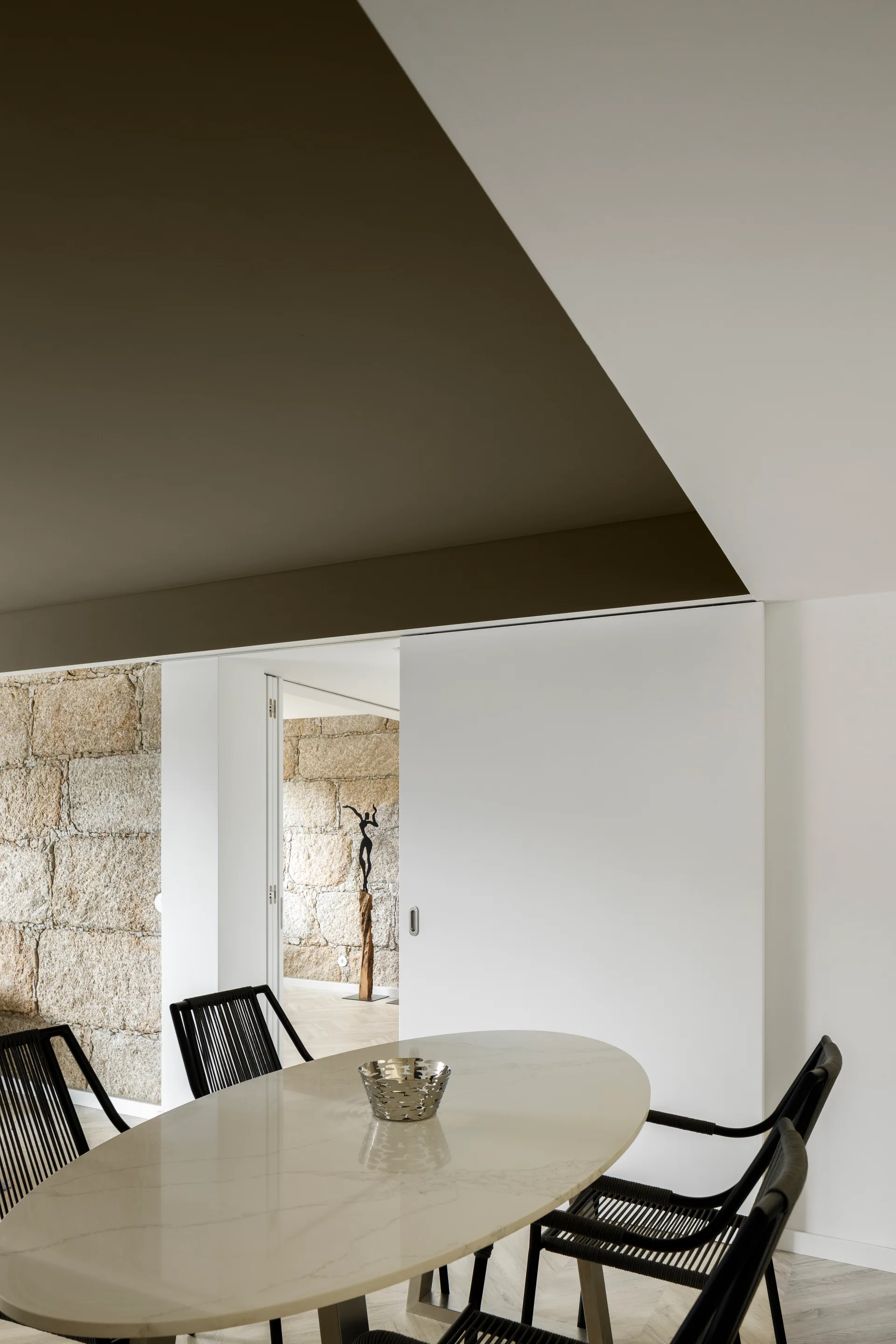 An oval marble dining table paired with black corded chairs is set against a rough stone wall and a large white sliding panel door.