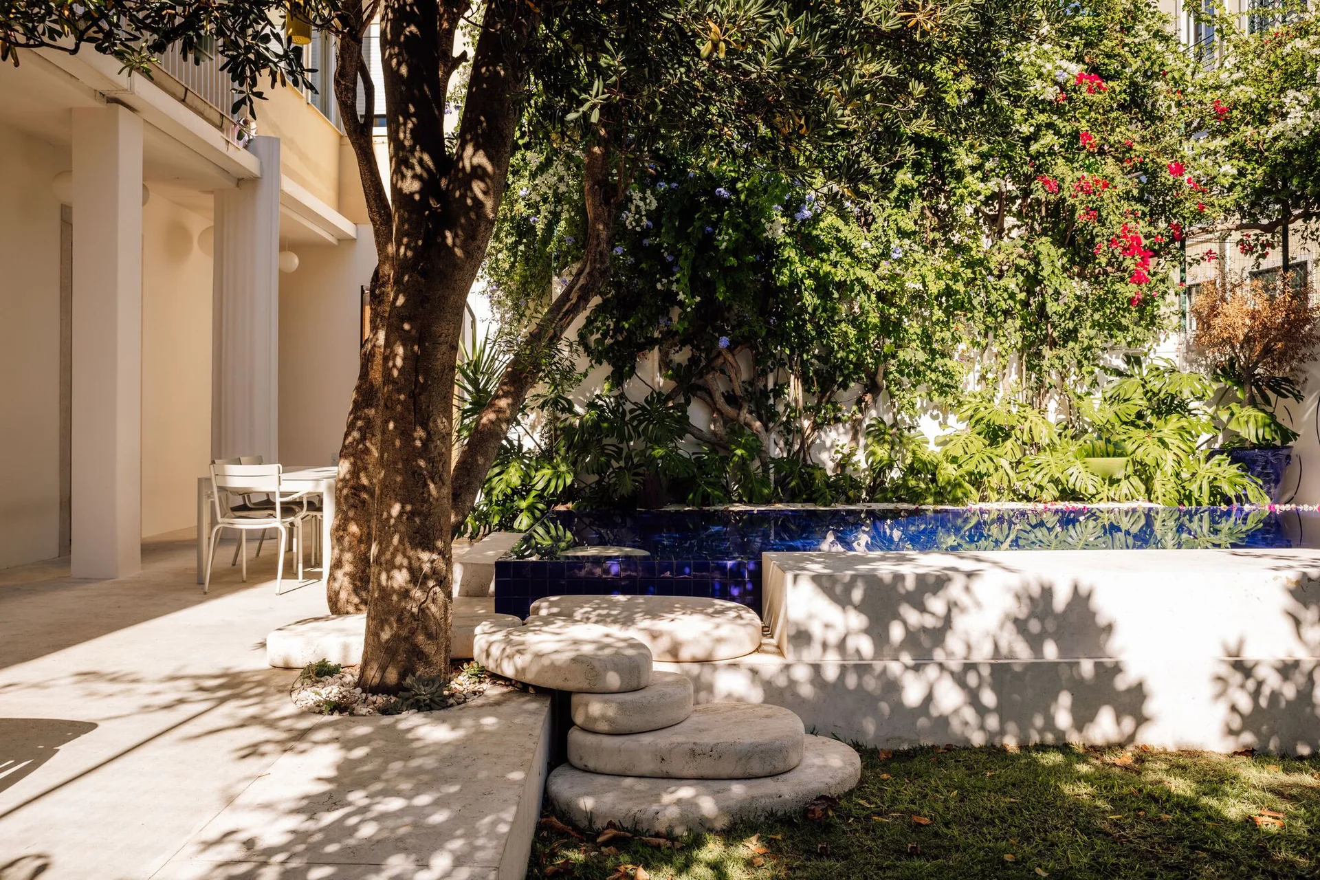 A sunlit courtyard featuring a blue tiled plunge pool, rounded stone steps wrapping around a tree, and a white outdoor dining table.