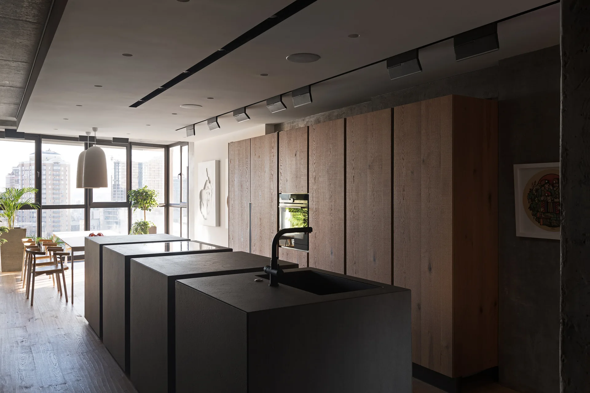 A minimalist kitchen features modular black geometric islands, a large wall of oak wood cabinets, and large floor-to-ceiling windows in the background.