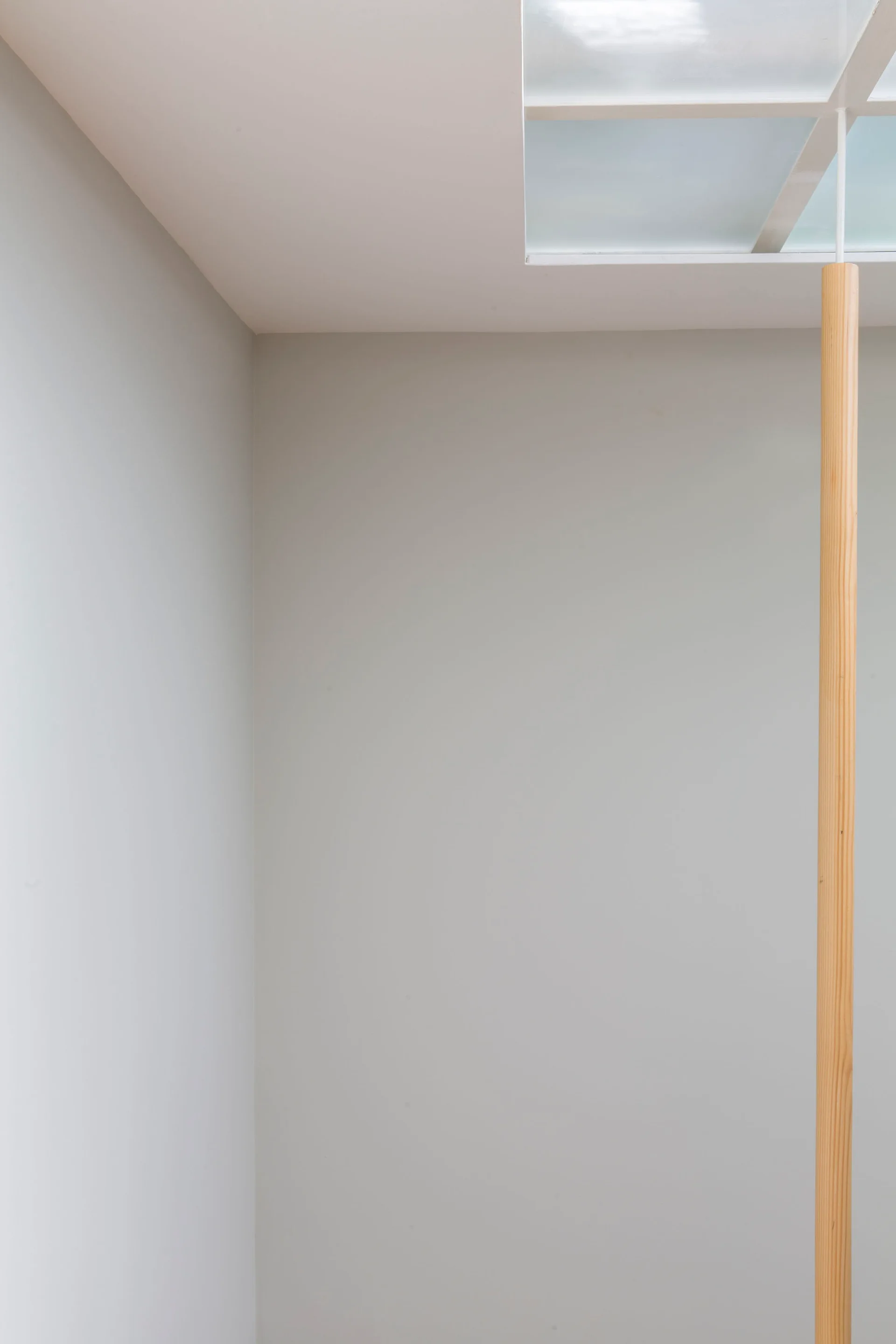 A minimalist light gray wall corner with a ceiling skylight supported by a single vertical pale wood post.