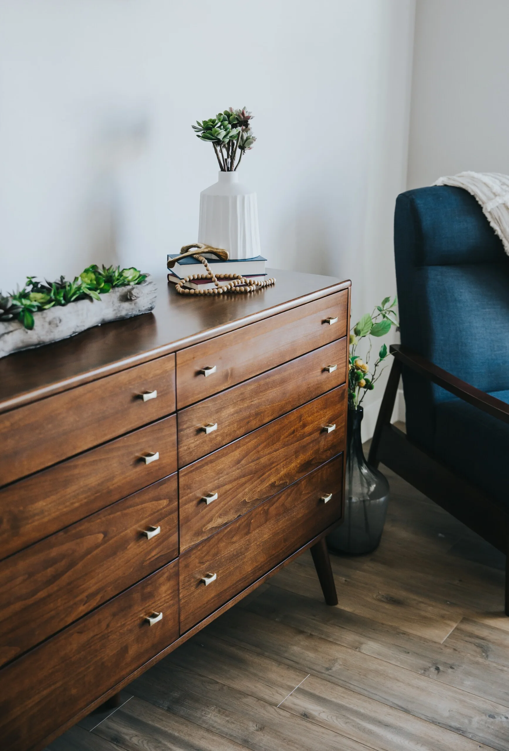 A mid-century modern wood dresser with metal drawer pulls sits on wood flooring next to a blue upholstered armchair in front of a white wall.