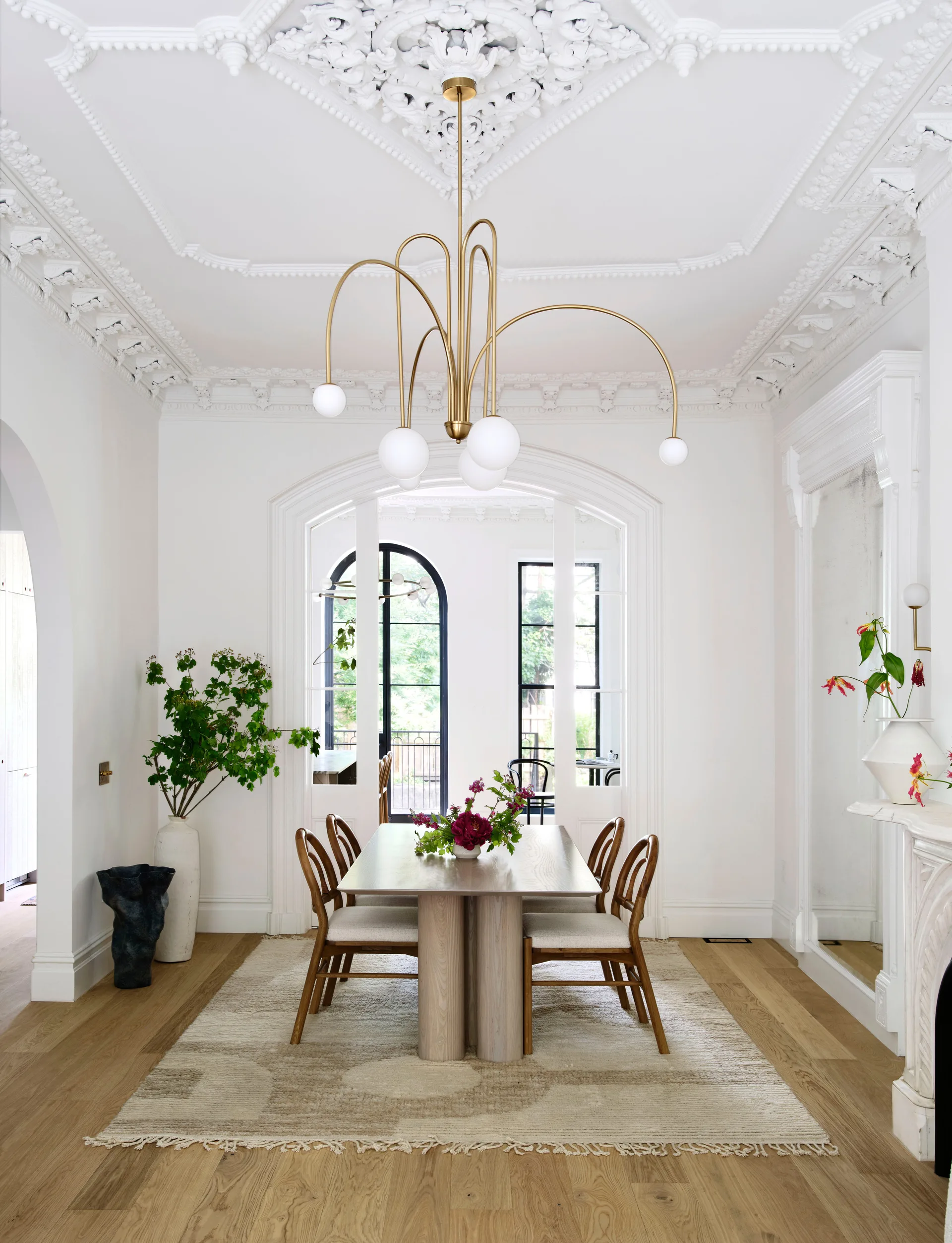 A light-filled dining room features a modern wood table and chairs under an ornate plaster ceiling with a brass globe chandelier.