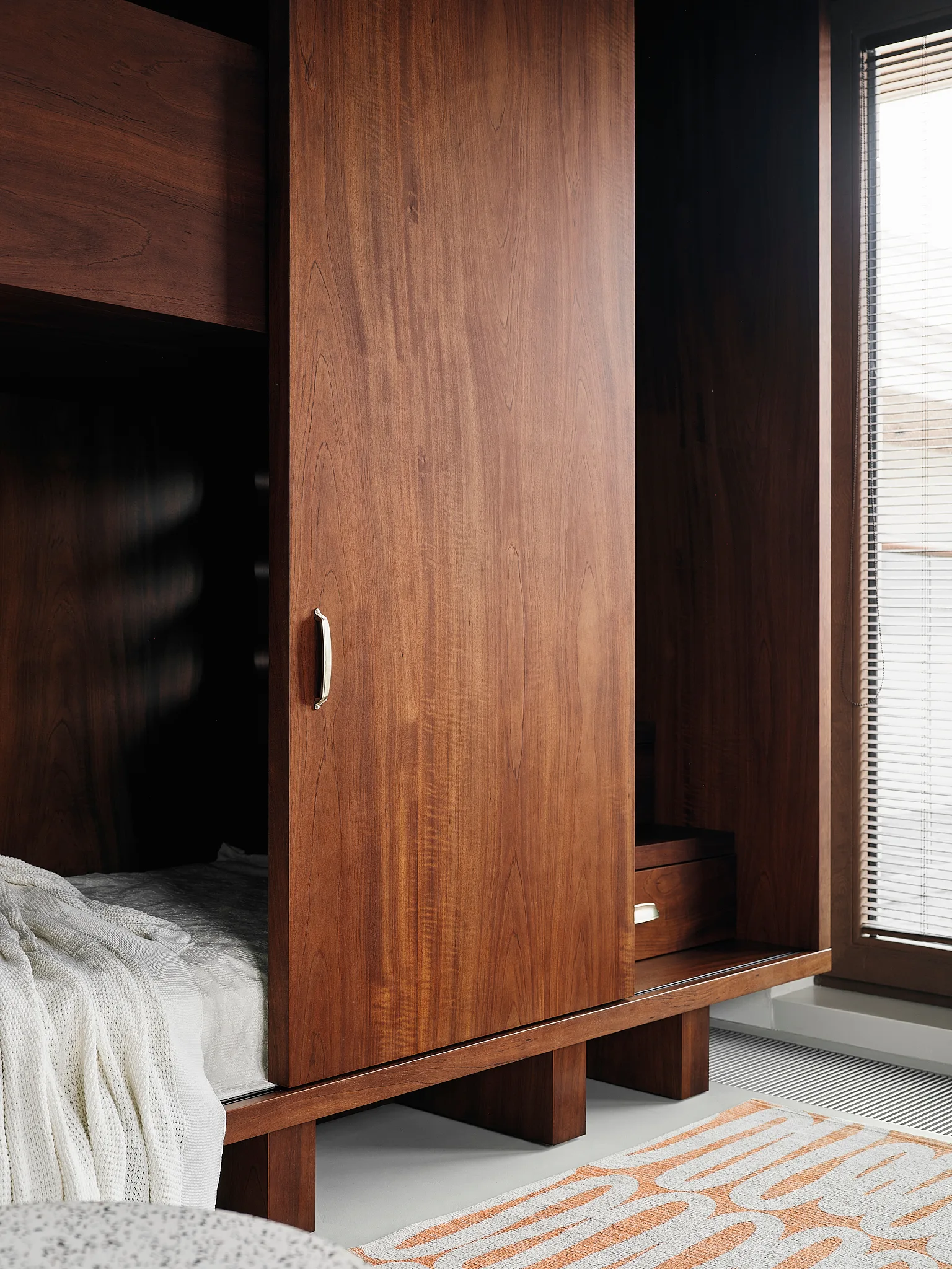 A close-up of a warm wood built-in wardrobe with a sliding door next to a bed with a white throw and a geometric patterned rug.