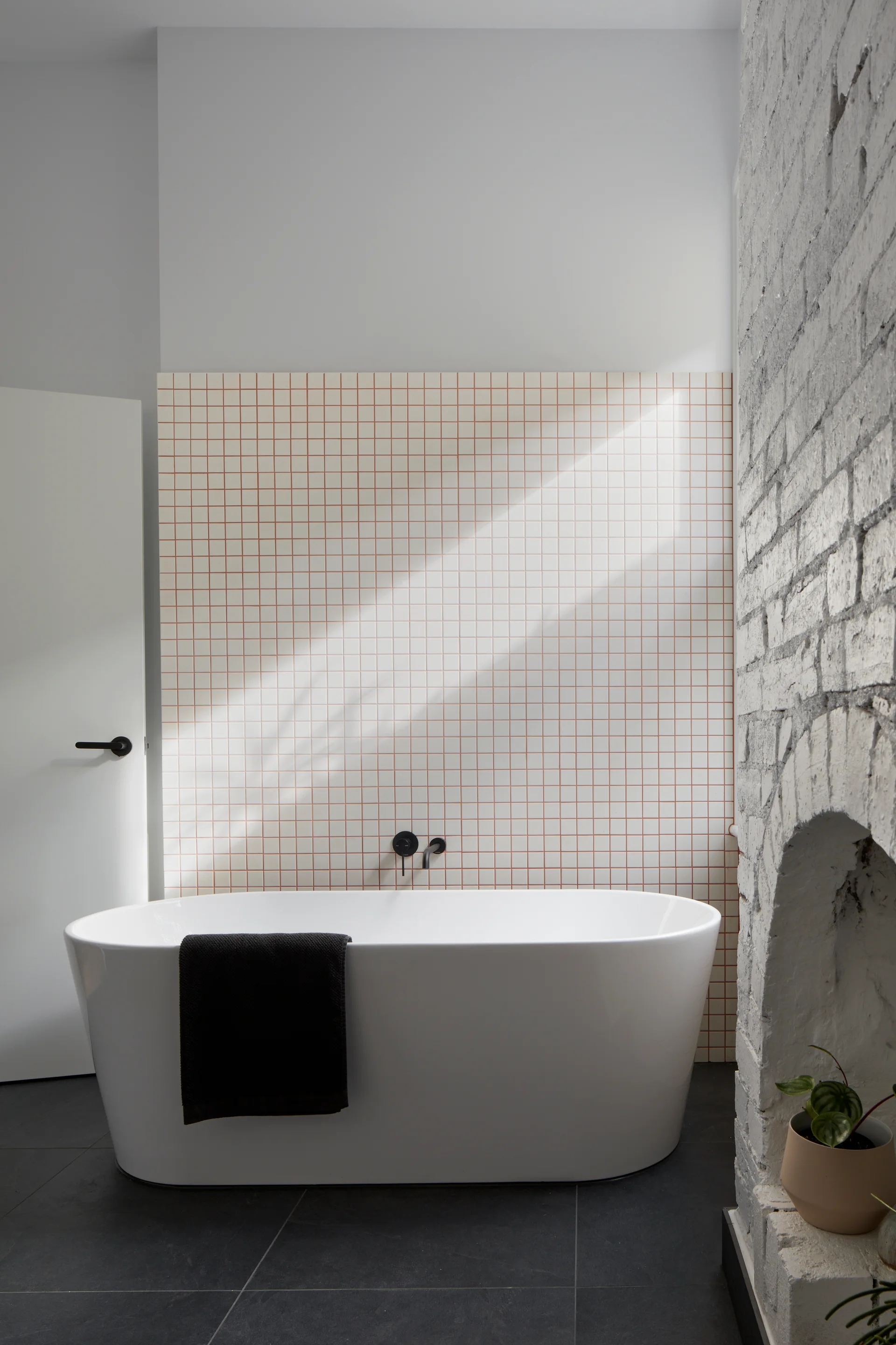 A white freestanding bathtub sits on dark floor tiles in front of a square grid tile wall and a white painted brick archway.