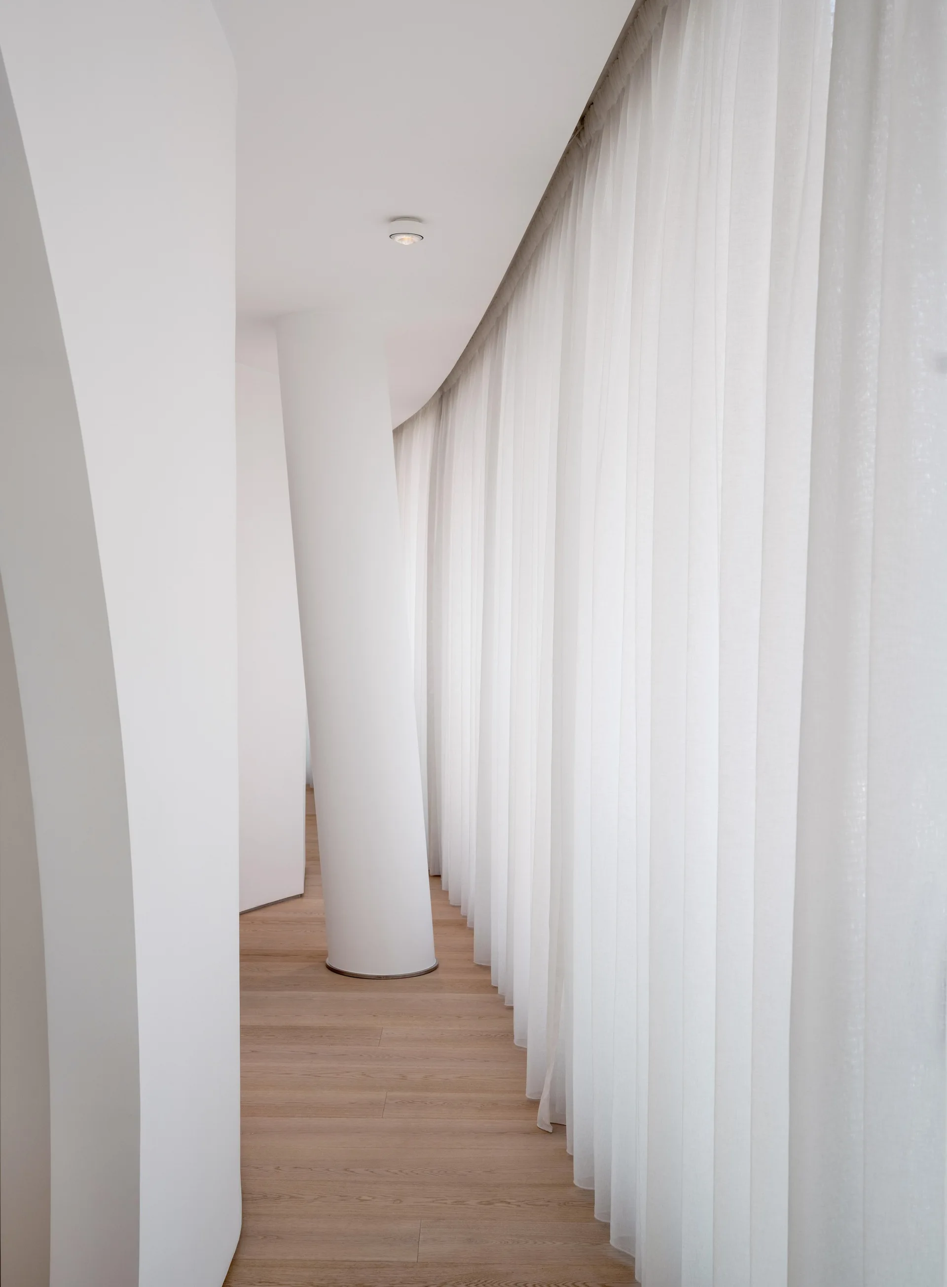A minimalist white corridor features light wood flooring, a central structural column, and floor-to-ceiling sheer white curtains along a curved wall.