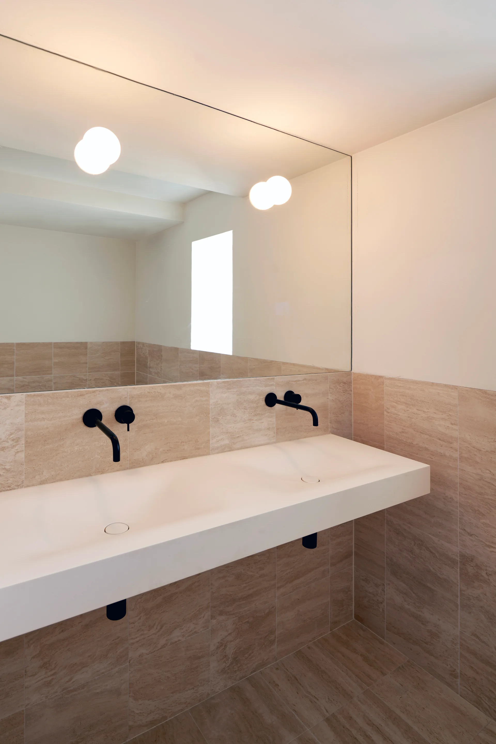 A modern bathroom features a floating white double sink with matte black wall-mounted faucets, beige wall tiles, and a large mirror.