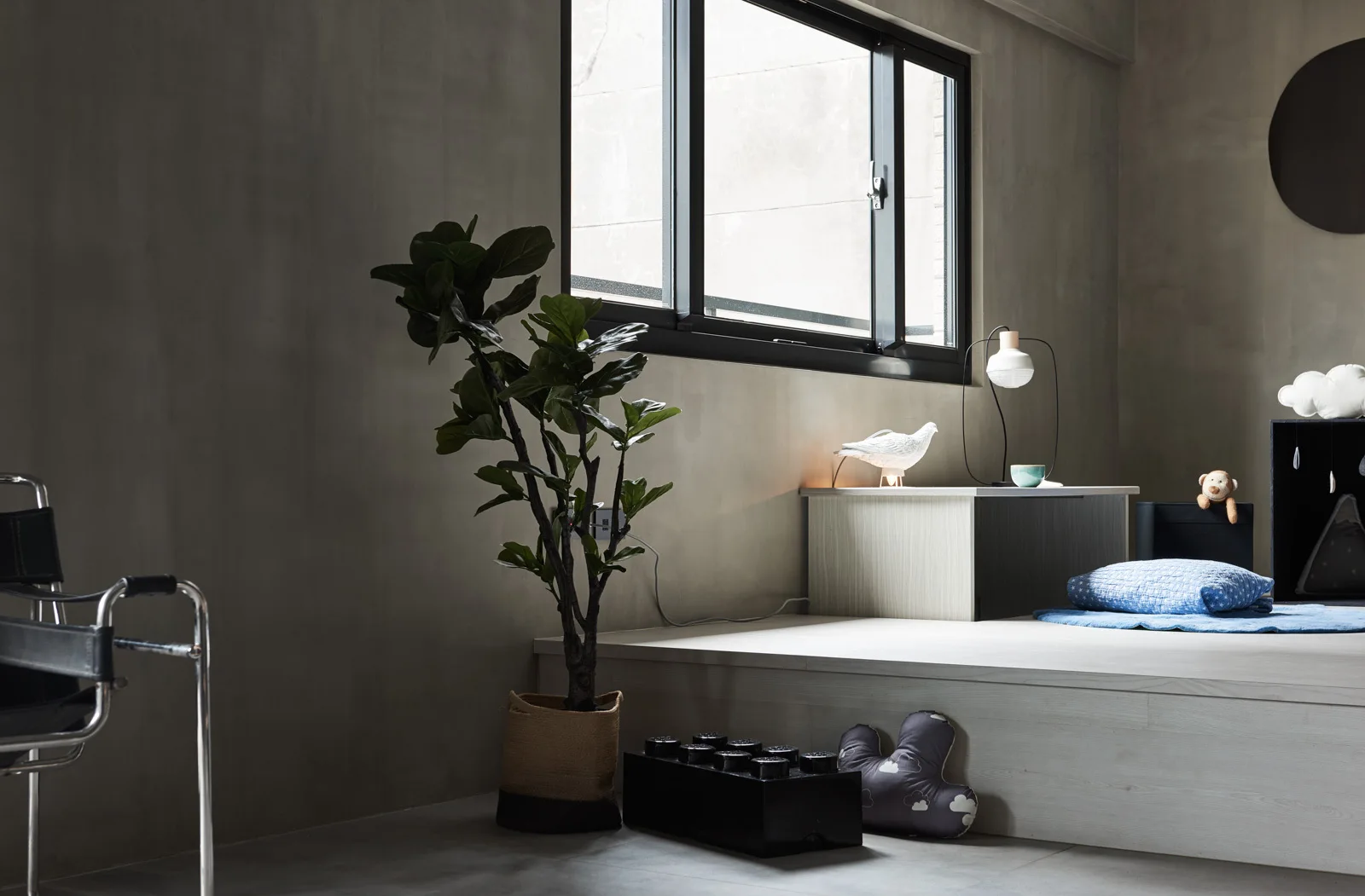 A minimalist children's bedroom features a raised wooden platform under a large window against grey concrete walls, decorated with a potted plant and toys.