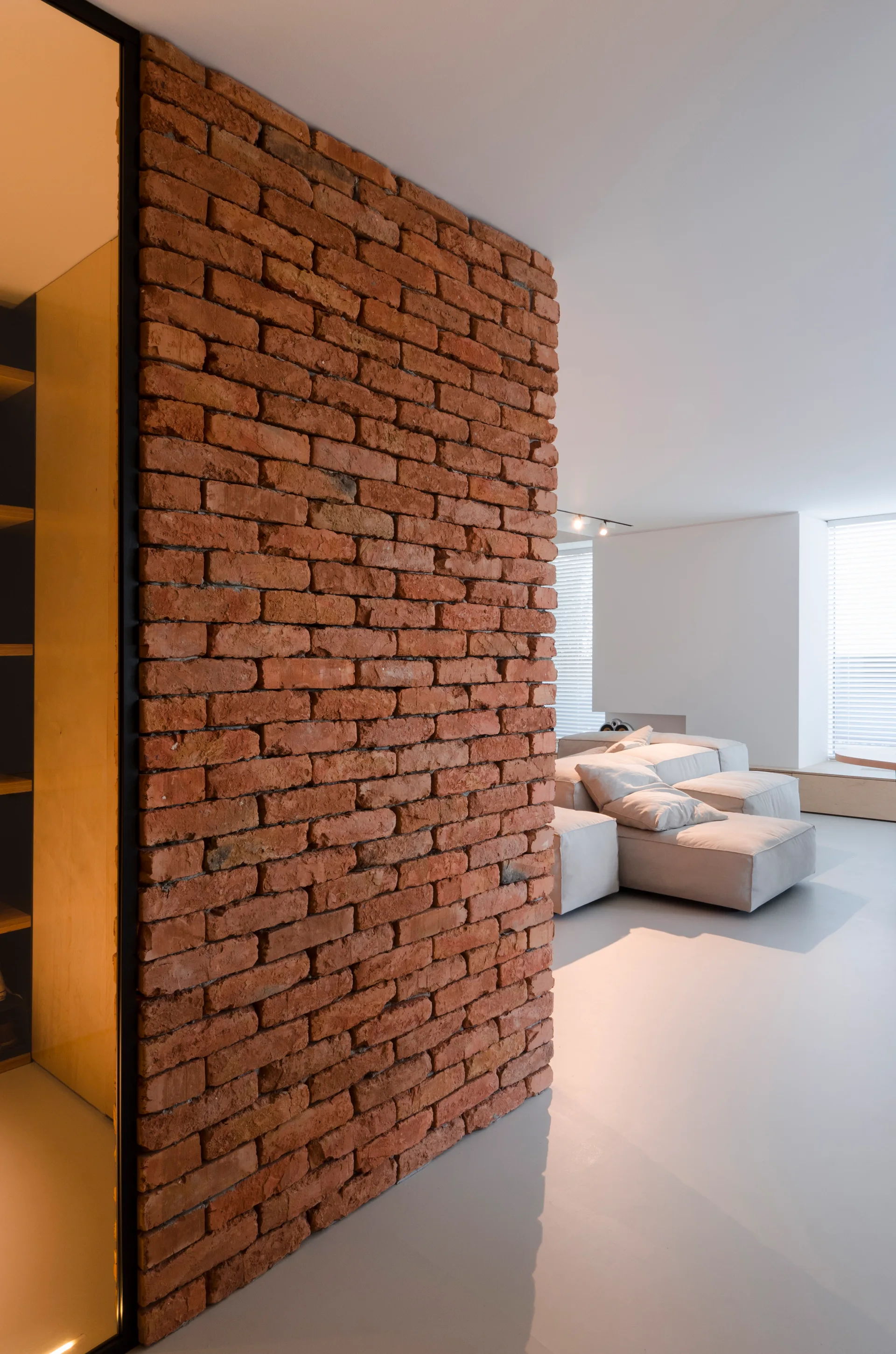 A minimalist living room featuring a prominent red brick partition wall, a low-profile light grey modular sofa, and smooth grey floors.
