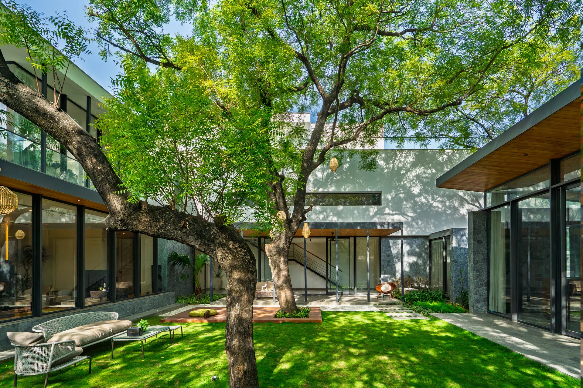 A modern residential courtyard features glass-walled wings wrapping around a green lawn with a large mature tree, wood decking, and outdoor seating.