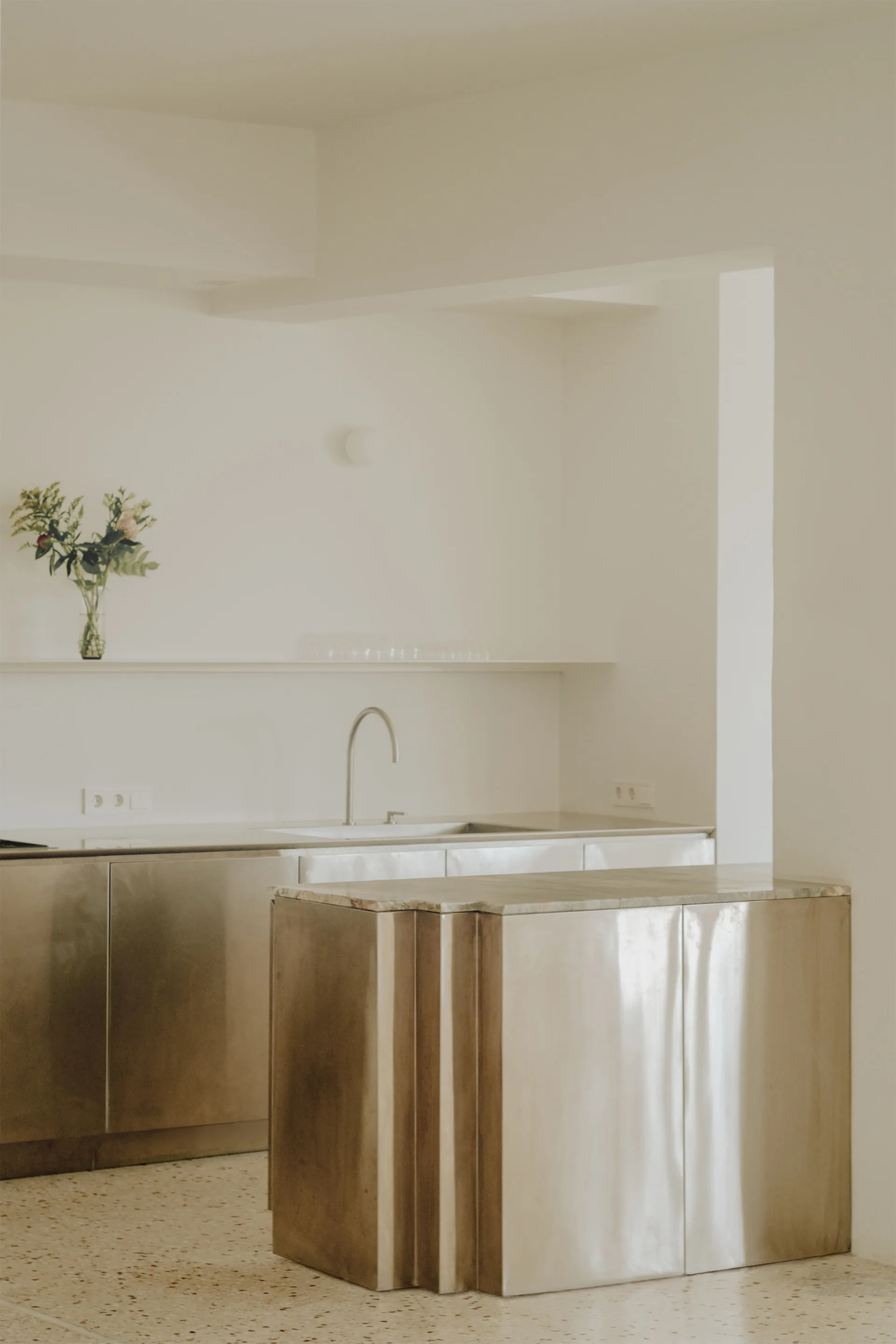 A minimalist kitchen features brushed stainless steel cabinetry, a matching metal island with a stone top, and beige terrazzo flooring under smooth white walls.