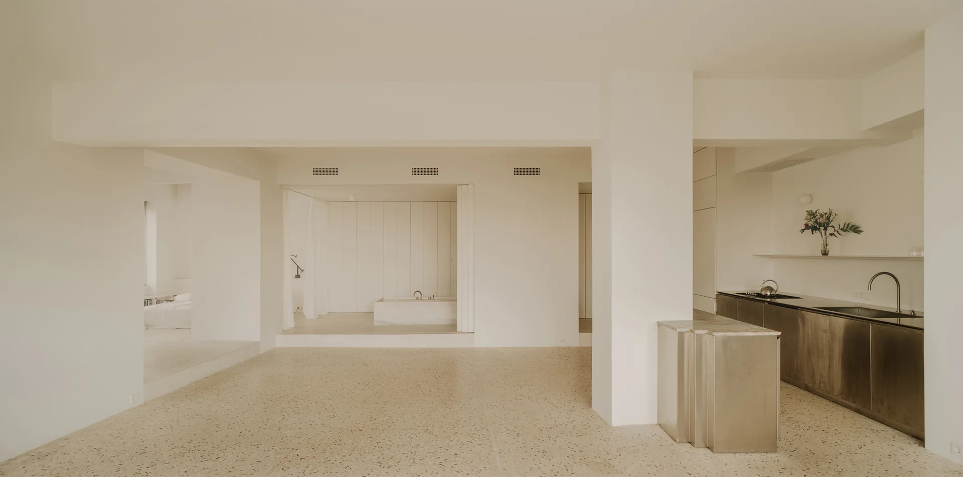 An open-concept minimalist room with light terrazzo flooring features a stainless steel kitchen counter, a raised bathroom alcove, and painted cream walls.