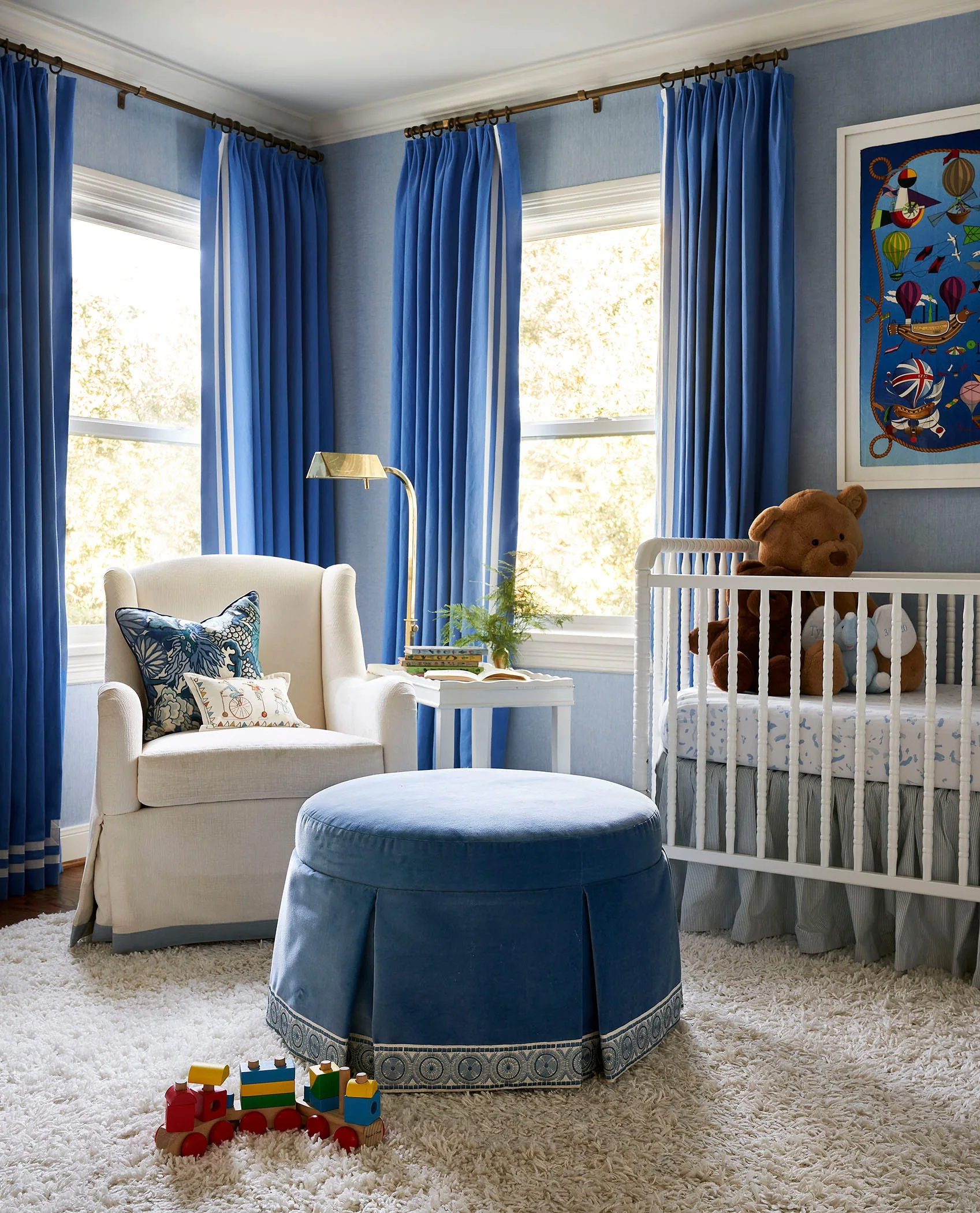 A blue nursery features a white crib, a cream armchair with a blue-trimmed skirt, a round blue pleated ottoman, and matching blue curtains.
