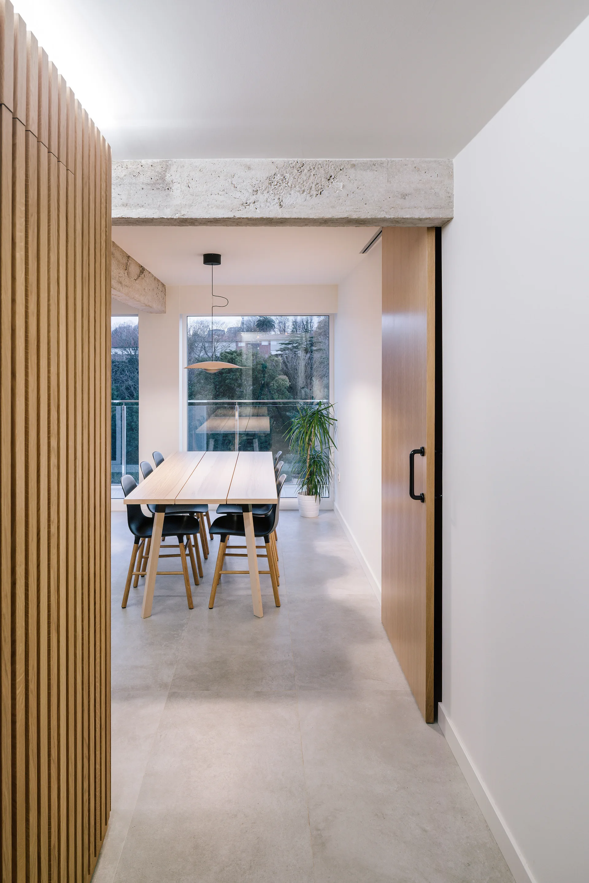 A modern dining room features a light wood table with black chairs beneath a raw concrete beam, next to a wood-slatted wall and sliding door.