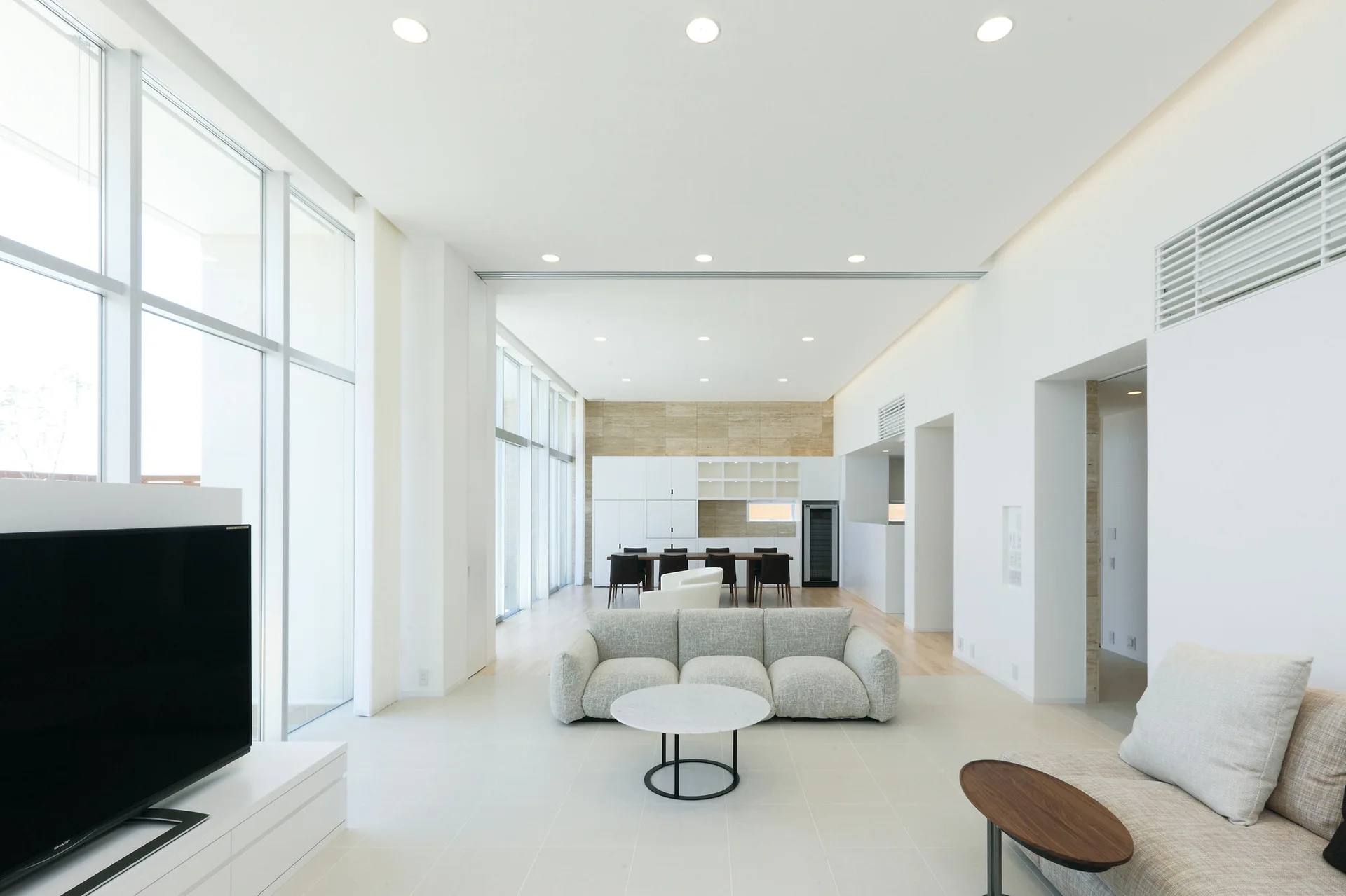 A minimalist open-concept living room features a low-profile grey sofa, floor-to-ceiling windows, and a dining table in the background.