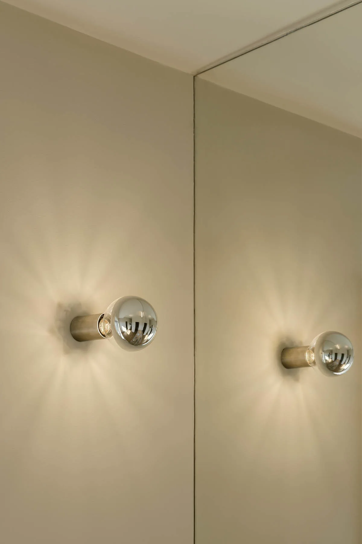 A warm-glowing chrome globe sconce on a beige painted wall is reflected in an adjacent frameless mirror.