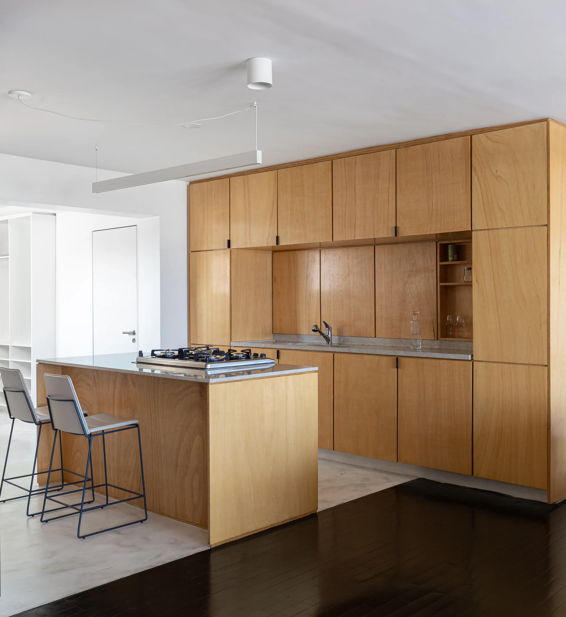 A minimalist kitchen features full-height plywood cabinets, a matching wood island with a stone countertop, and two-toned flooring.