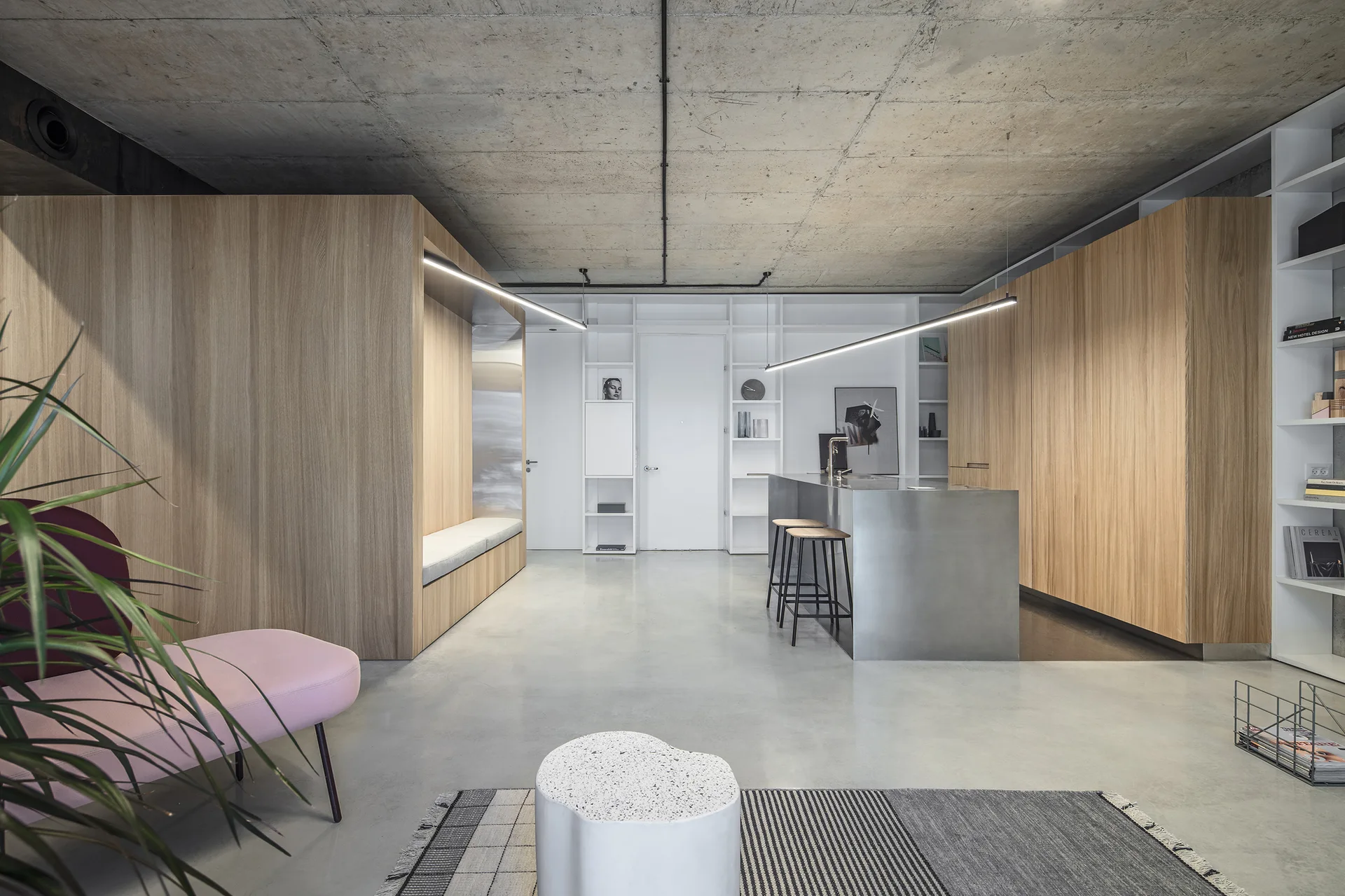 A minimalist kitchen features a brushed metal island, wood-paneled built-ins, a built-in bench, and an exposed concrete ceiling with linear lights.