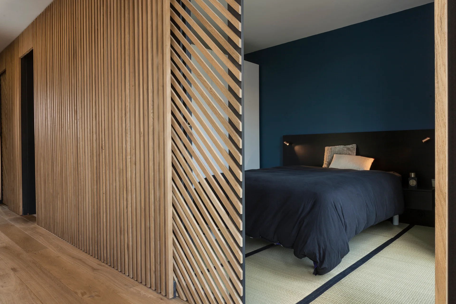 A modern bedroom features a navy blue wall behind a dark bed, seen through a sliding wooden partition with vertical and diagonal slats.