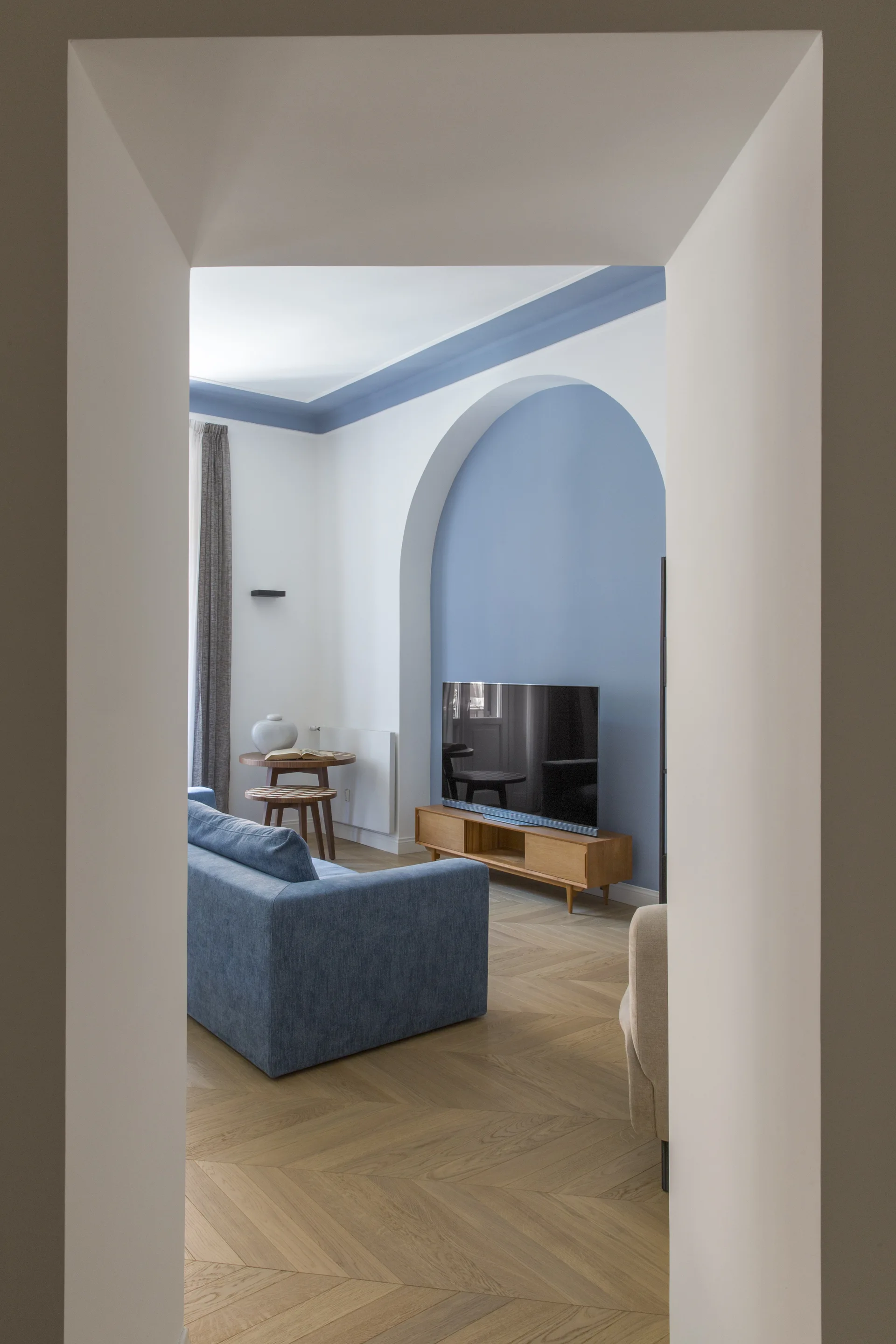 Viewed through a doorway, a living room features a blue fabric sofa, herringbone wood floors, and a television resting in a blue arched wall alcove.
