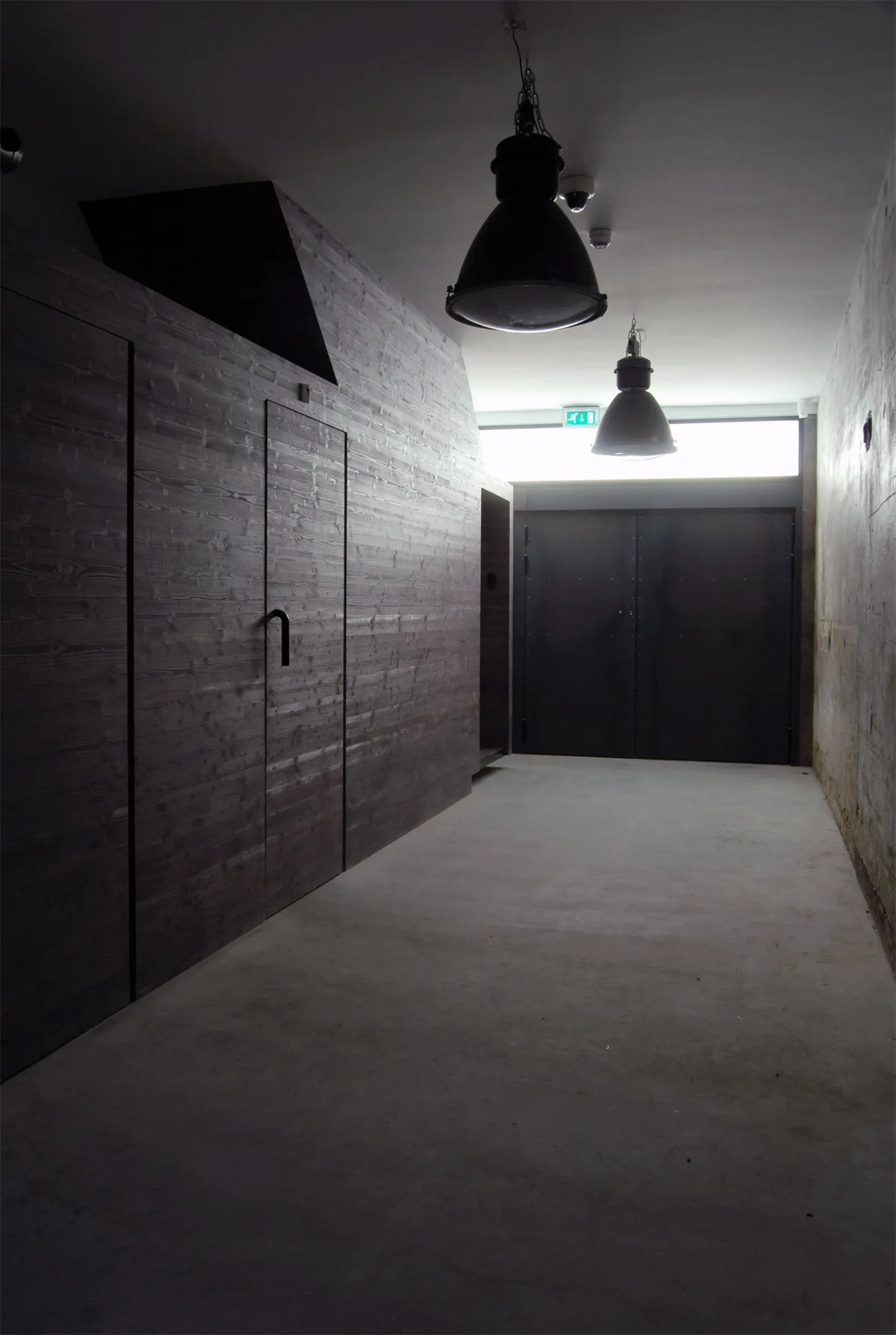 An industrial hallway features dark wood paneled walls, a raw concrete floor, and two large black pendant lights hanging from the ceiling.