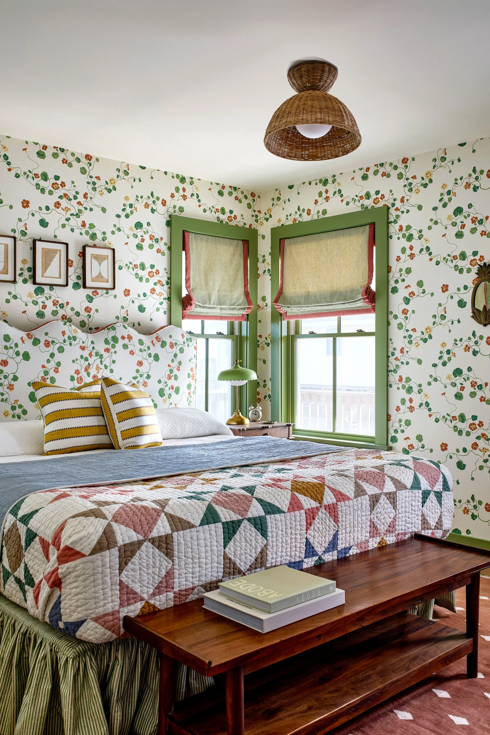A cozy bedroom featuring nasturtium floral wallpaper, matching green window trim, a scalloped headboard, and a patchwork quilt.