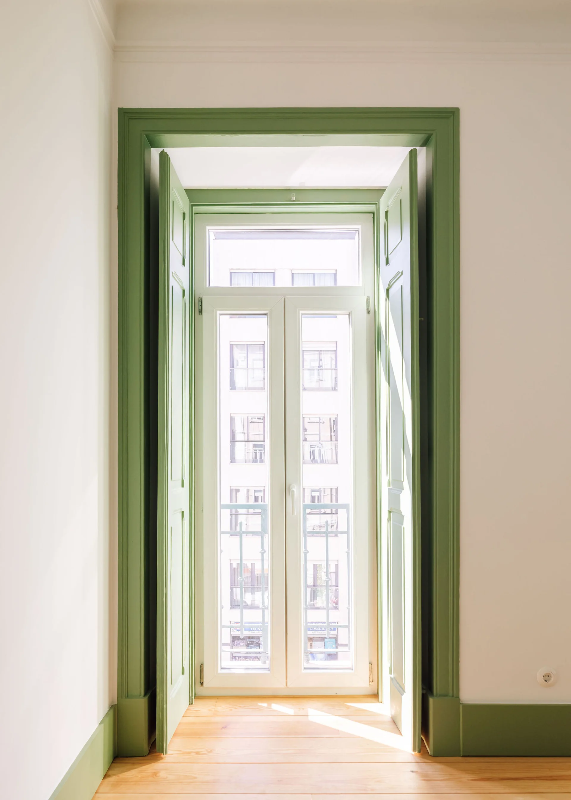 A tall white casement window with open sage green wooden folding shutters, matching green trim, white walls, and a light wood floor.