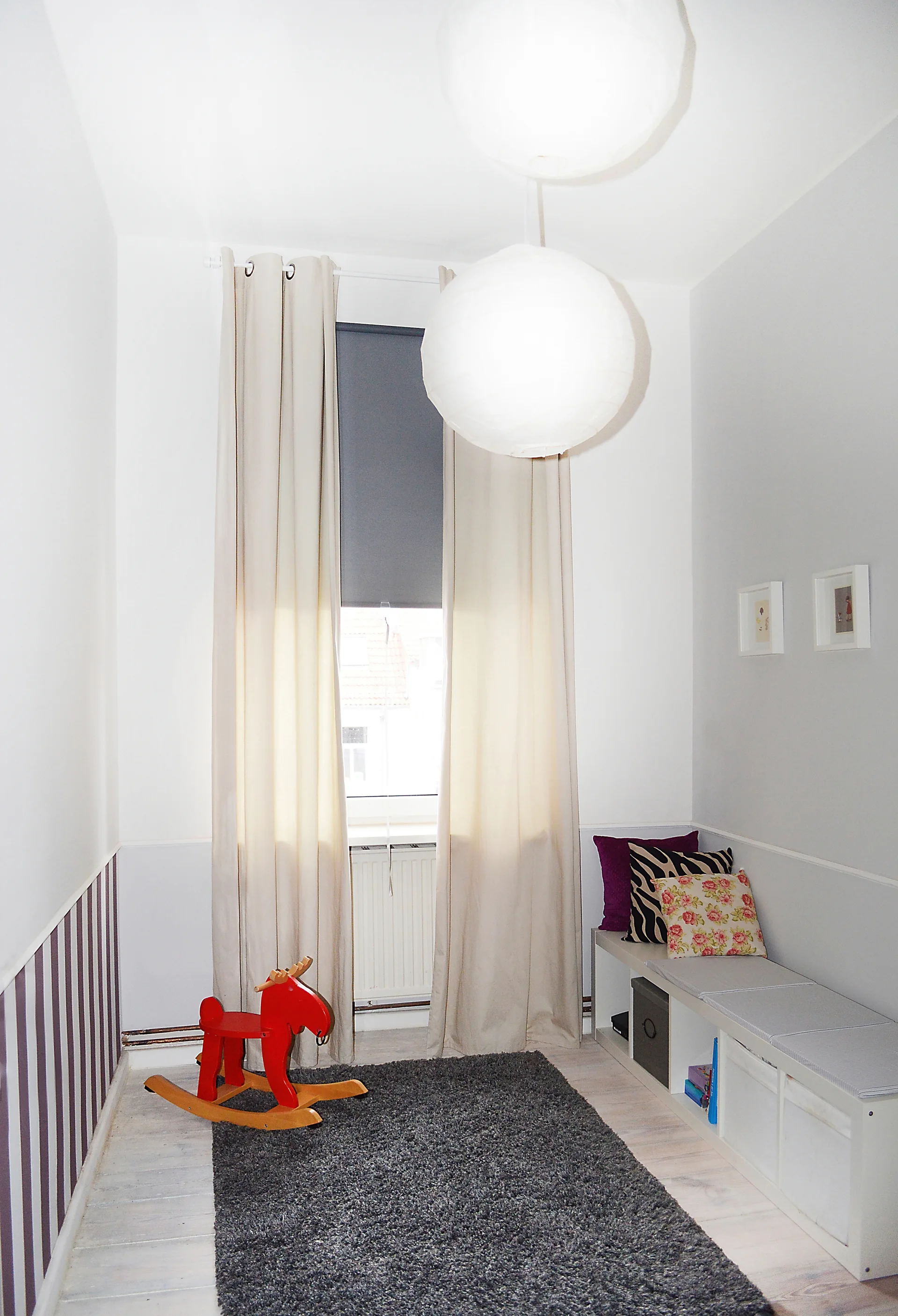 A small playroom features a long white storage bench with cushions, a red wooden rocking horse, and two paper pendant lights hanging from the ceiling.