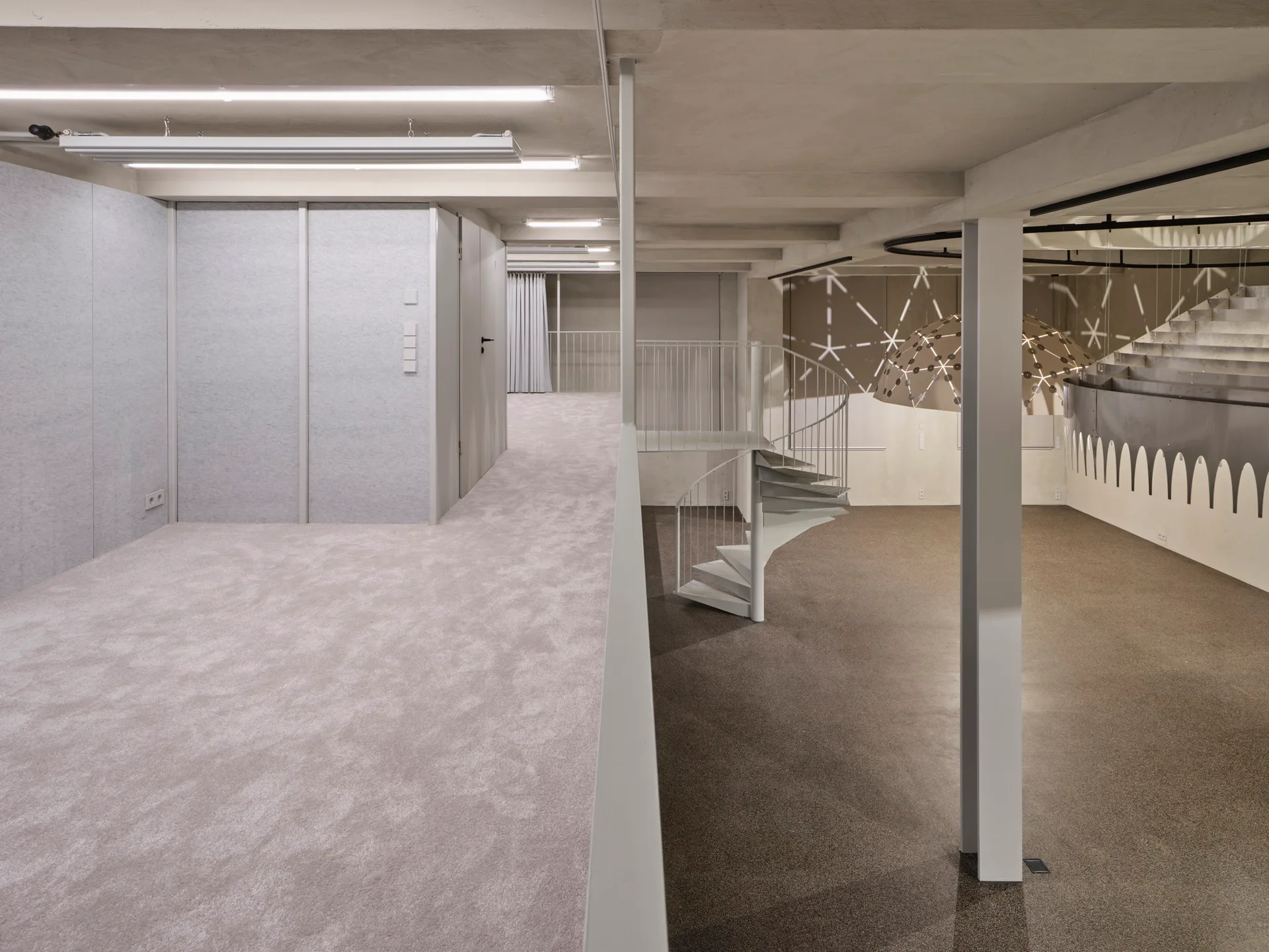 An open multi-level industrial space with a white metal spiral staircase, carpeted floors, exposed concrete ceilings, and a large dome pendant light.