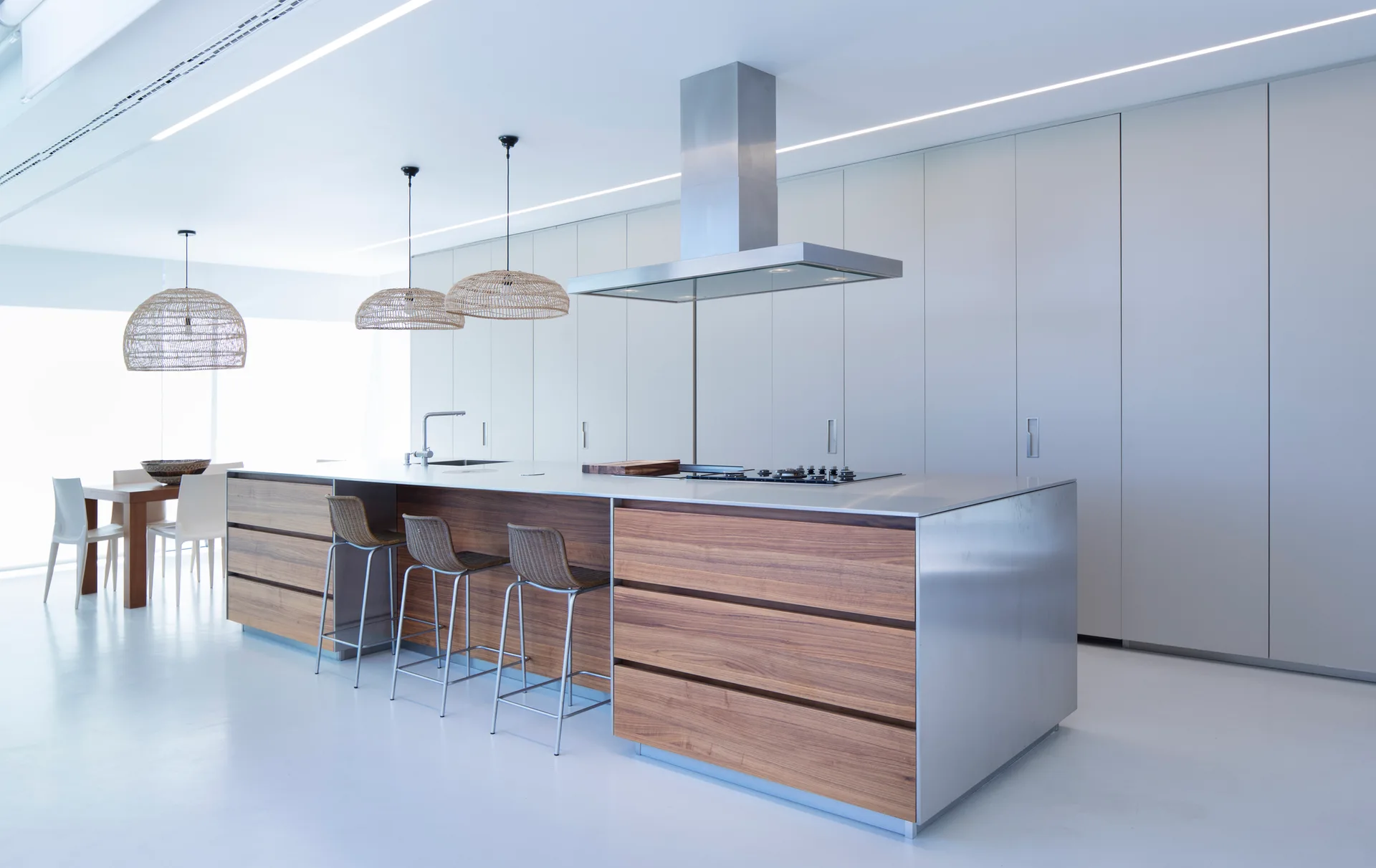 A minimalist kitchen features a long island with warm wood drawers and stainless steel details, under three woven dome pendant lights.