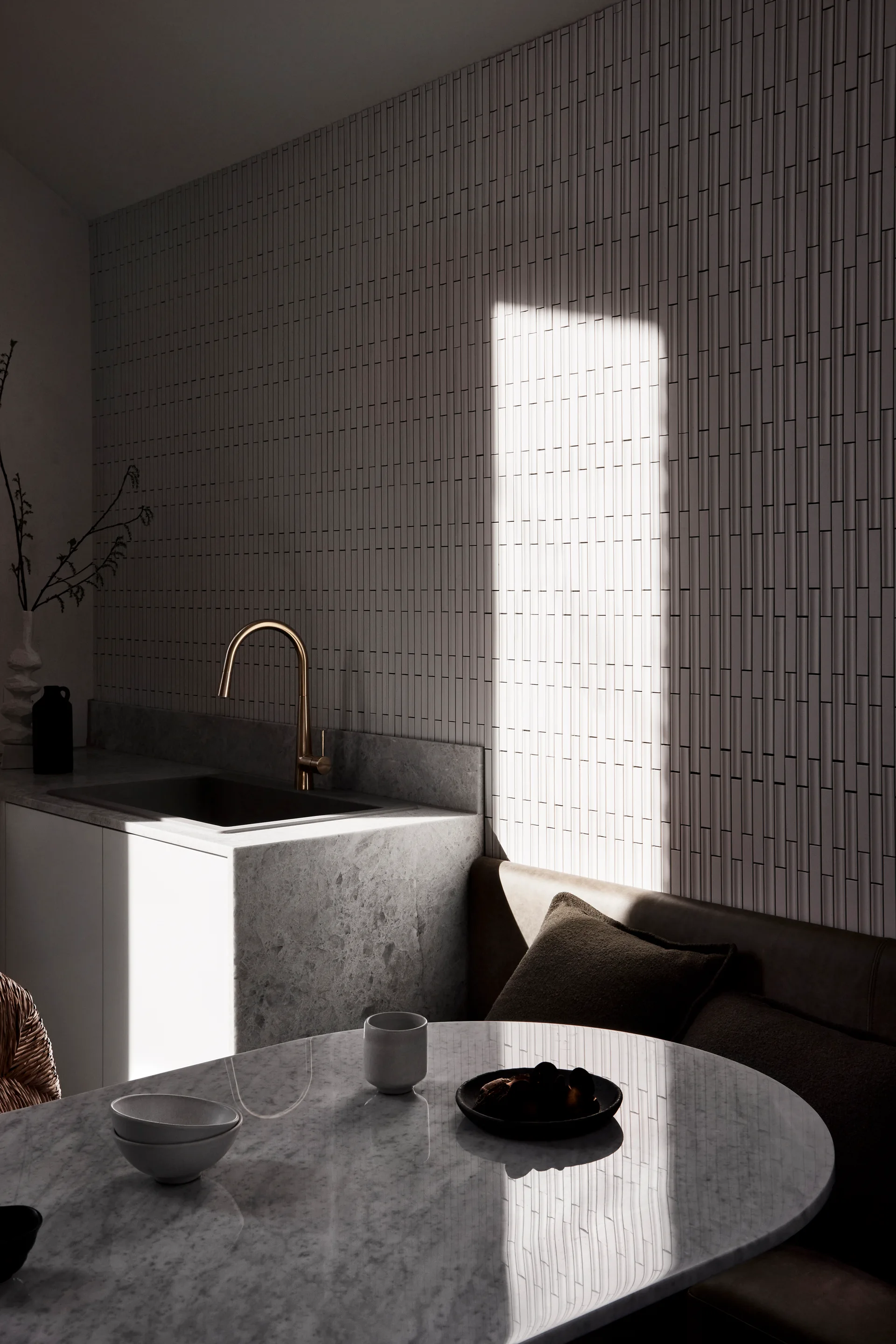 A minimalist kitchen corner features a vertical tiled wall, a marble sink counter with a brass faucet, and an oval marble dining table.