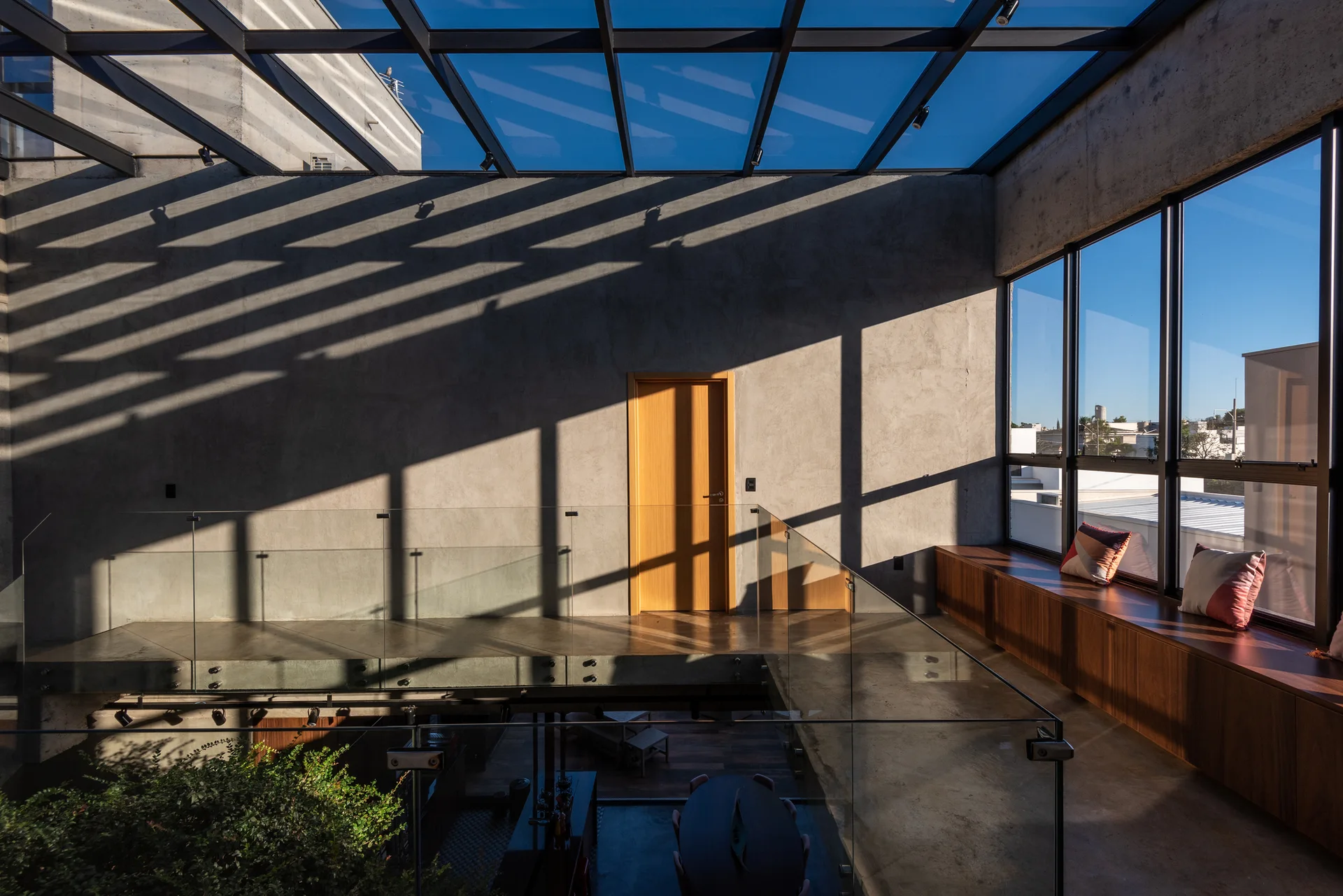 A modern landing featuring a concrete wall, glass roof skylight casting shadows, glass railings, and a wood-toned built-in storage bench.
