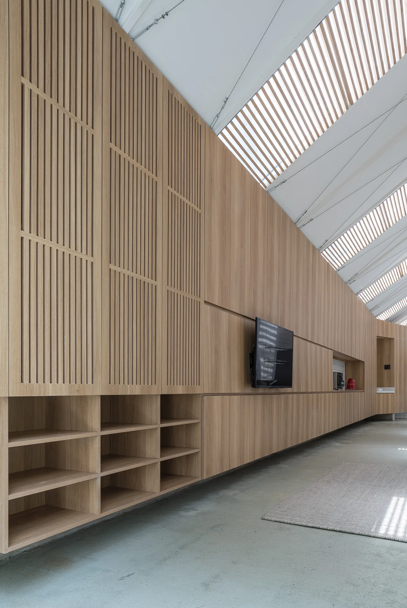 A floor-to-ceiling light wood cabinetry wall with slatted details and open shelves stands on a concrete floor beneath an angled ceiling with skylights.