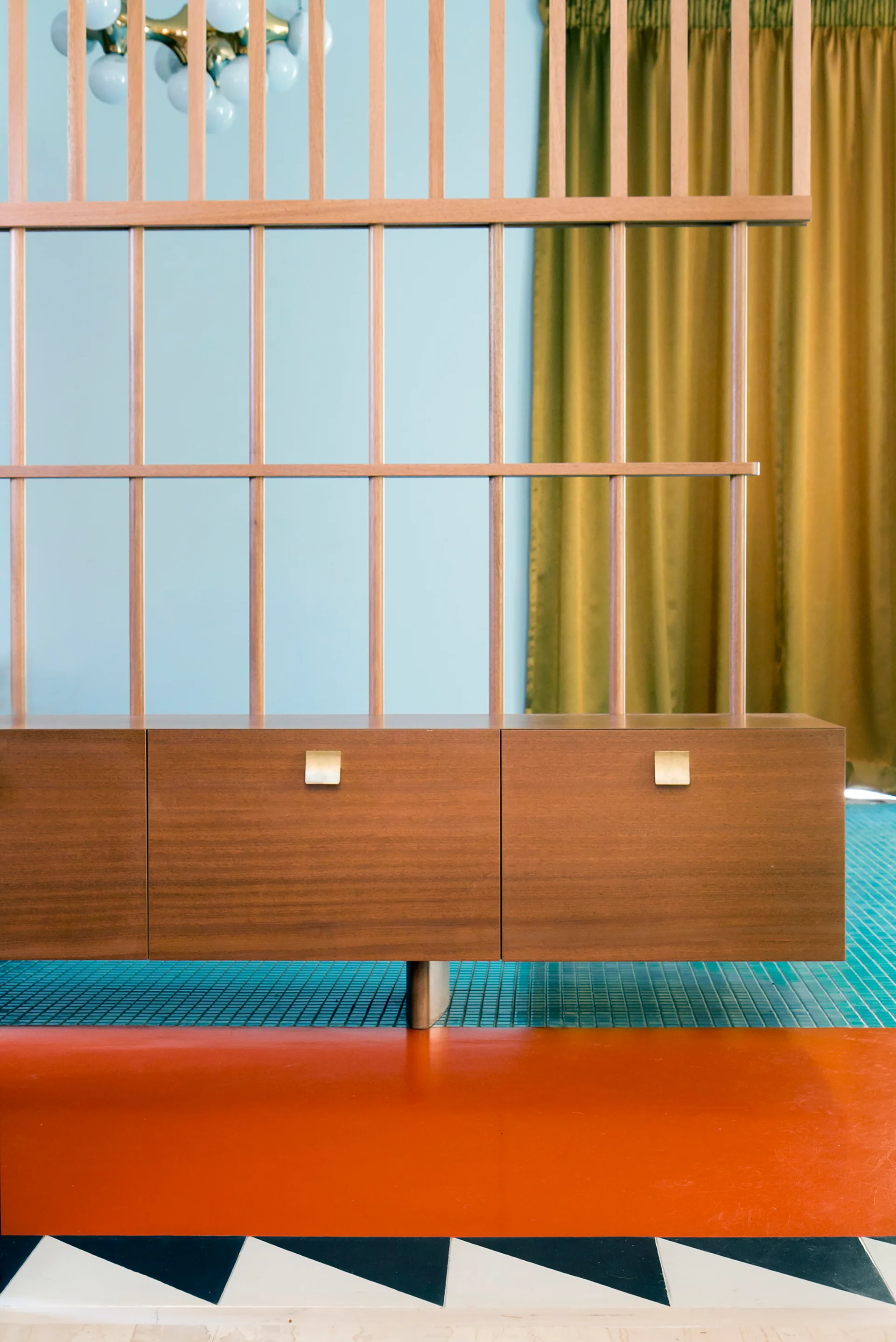 A wooden slatted room divider stands above a floating wood cabinet with gold pulls, set against a light blue wall and a colorful tiled floor.