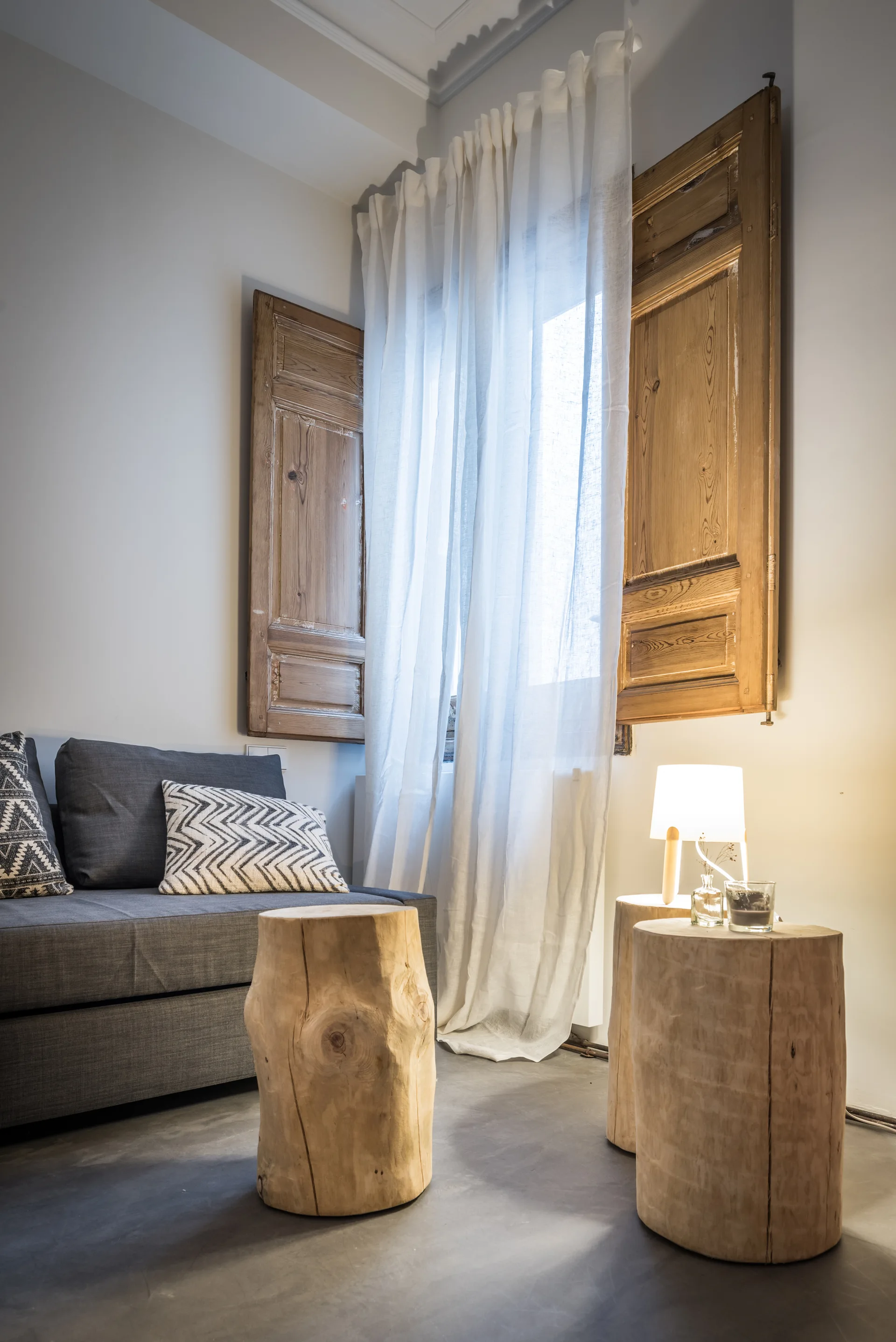 A grey sofa with patterned pillows sits next to natural tree stump side tables and a window with rustic wood shutters.