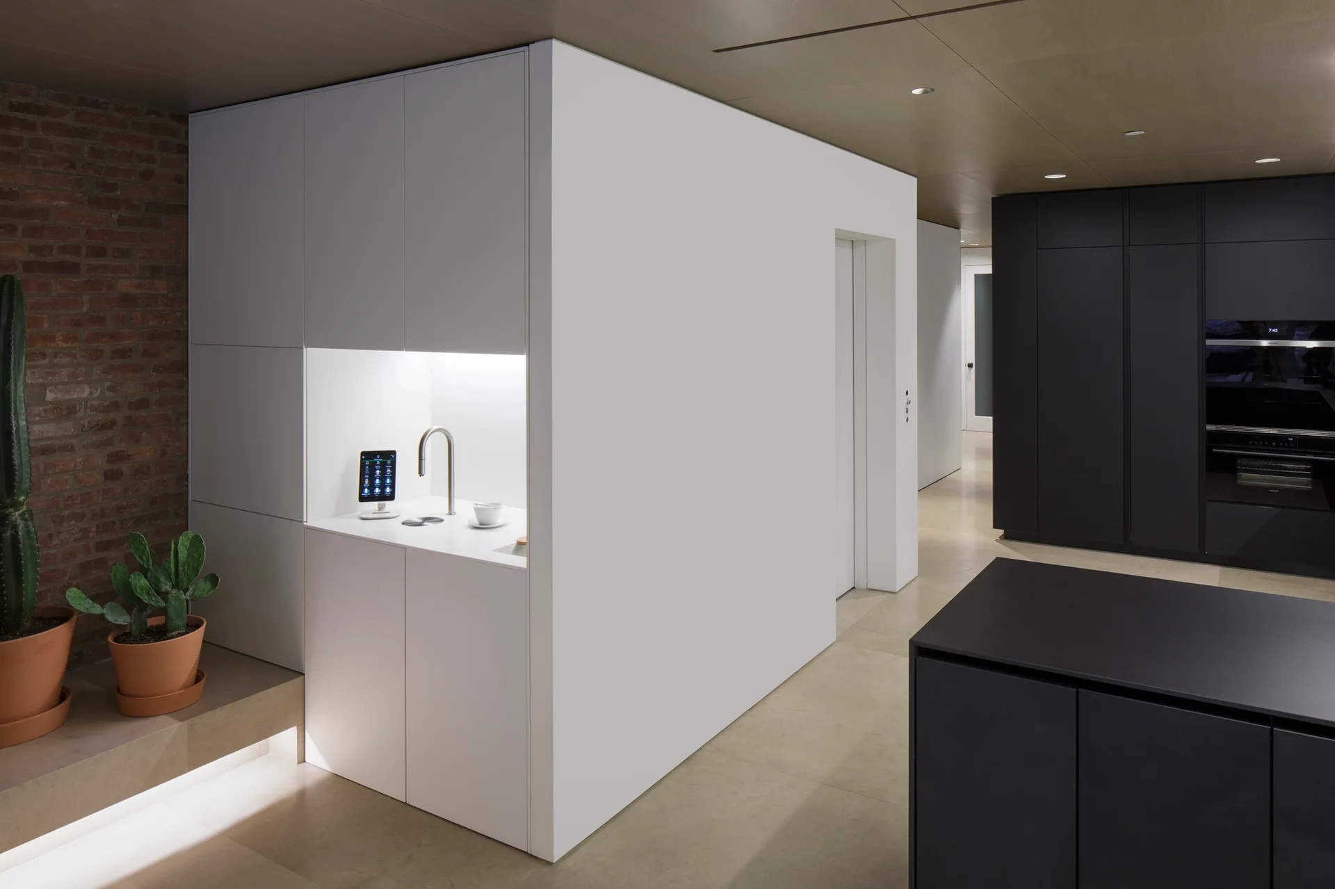 A modern kitchen featuring a monolithic white cabinet block with a built-in sink niche, matte black cabinets, wood ceiling, and a brick accent wall.