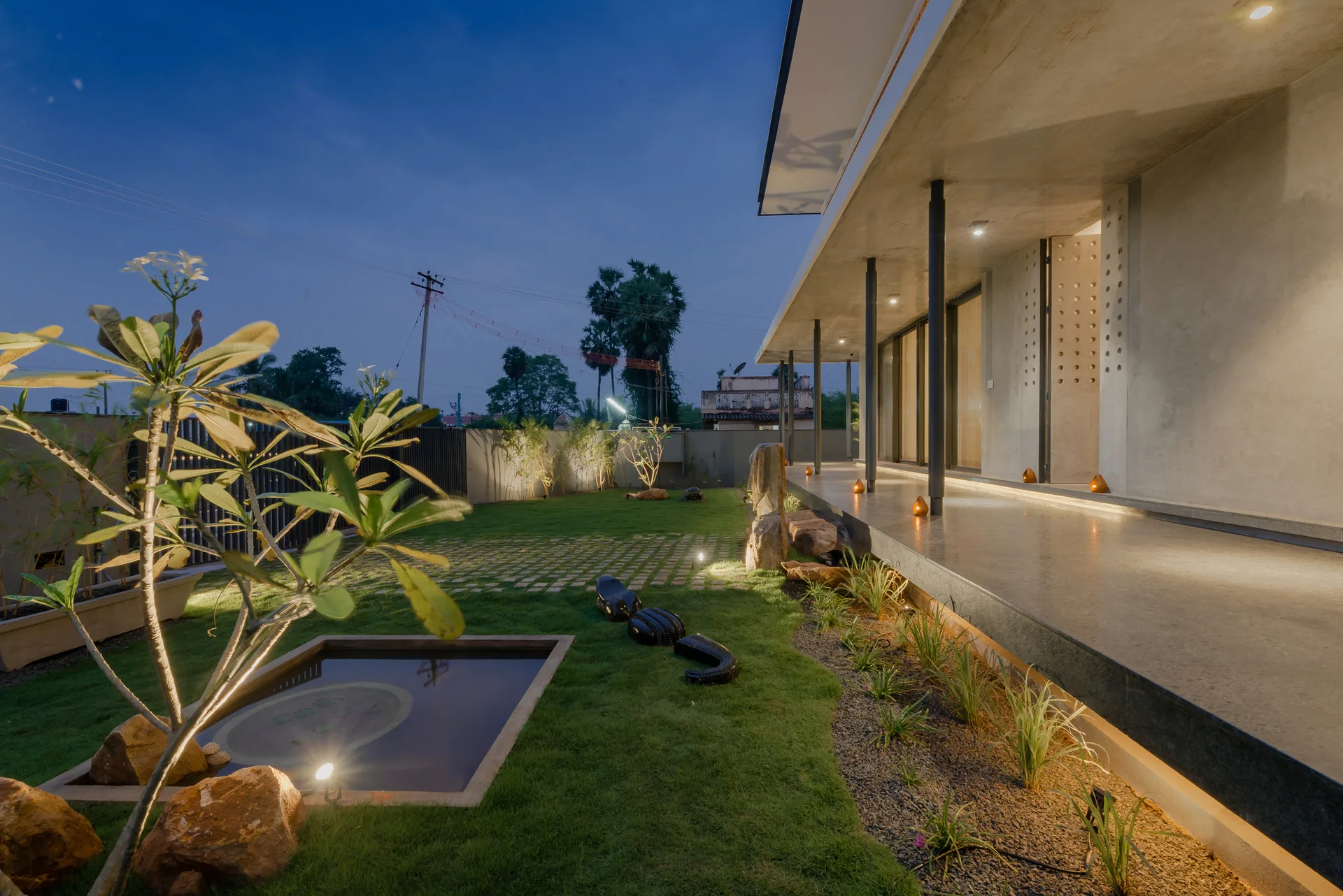 A modern concrete patio with columns and warm ground lighting overlooks a green grass lawn with a small square pond at dusk.