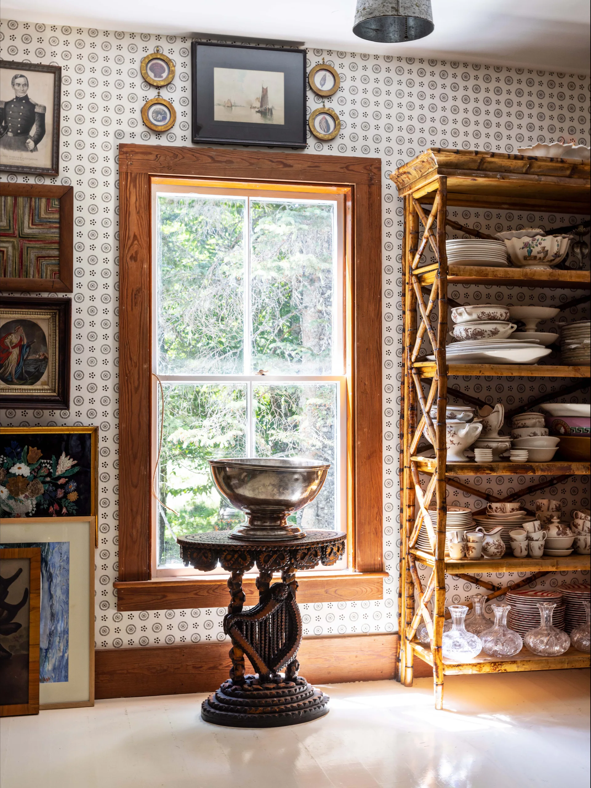 A bamboo etagere filled with ceramic dishware stands next to a window and carved wood table in a dining room with patterned wallpaper.