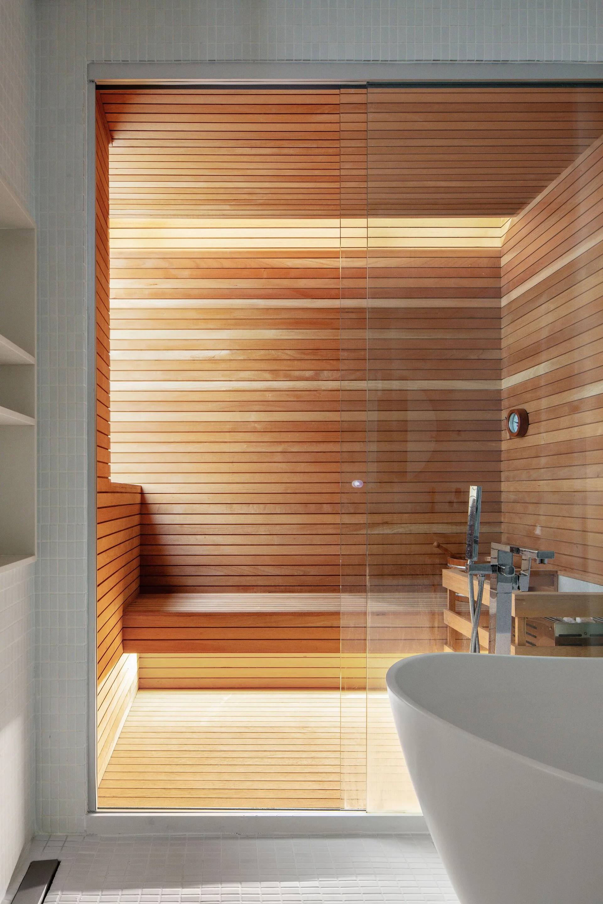 A modern bathroom featuring a glass-enclosed cedar wood sauna adjacent to a white freestanding bathtub on small square tiled flooring.