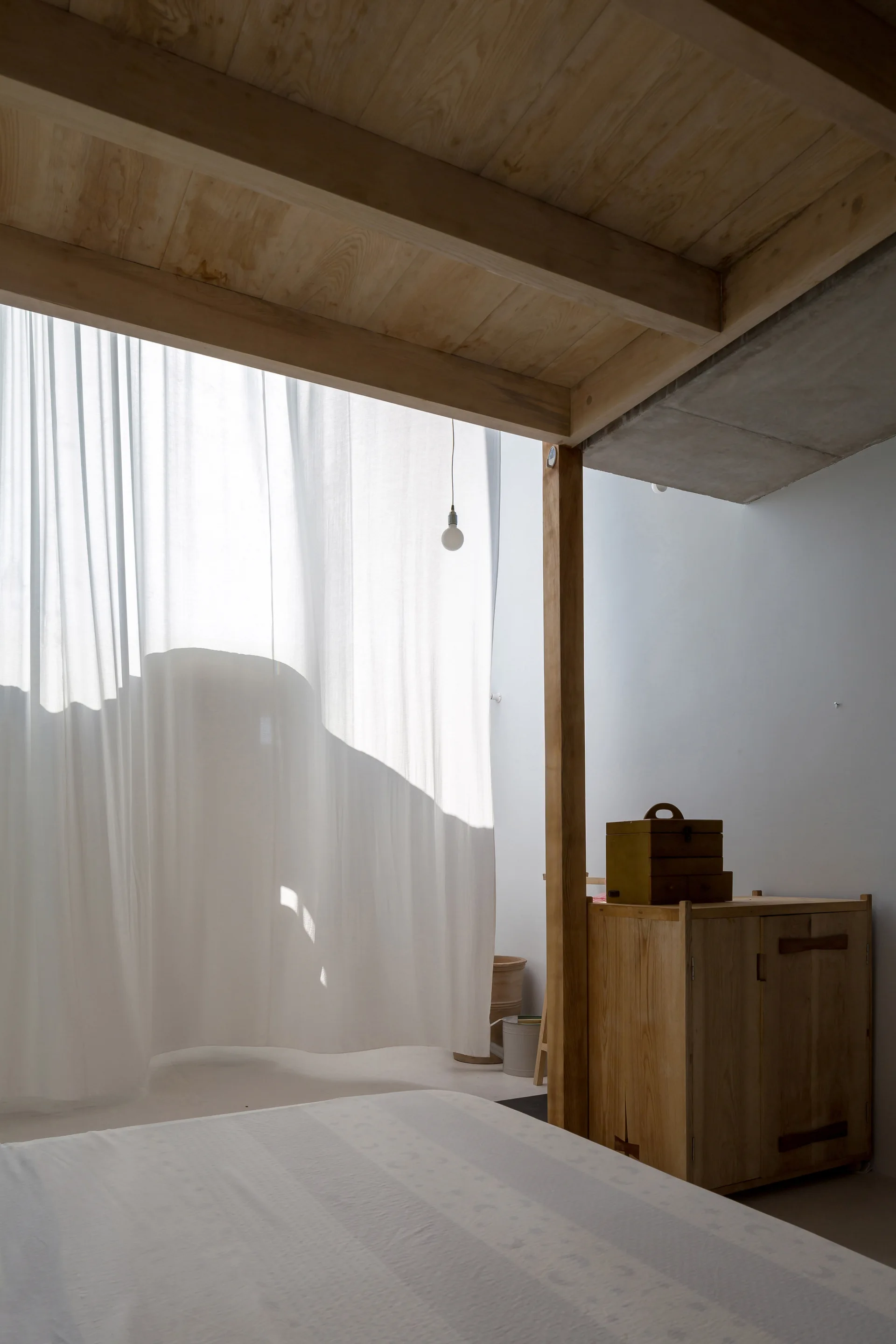 A minimalist bedroom features an exposed wood ceiling, a light wood storage cabinet, and a sheer white curtain filtering bright natural light.