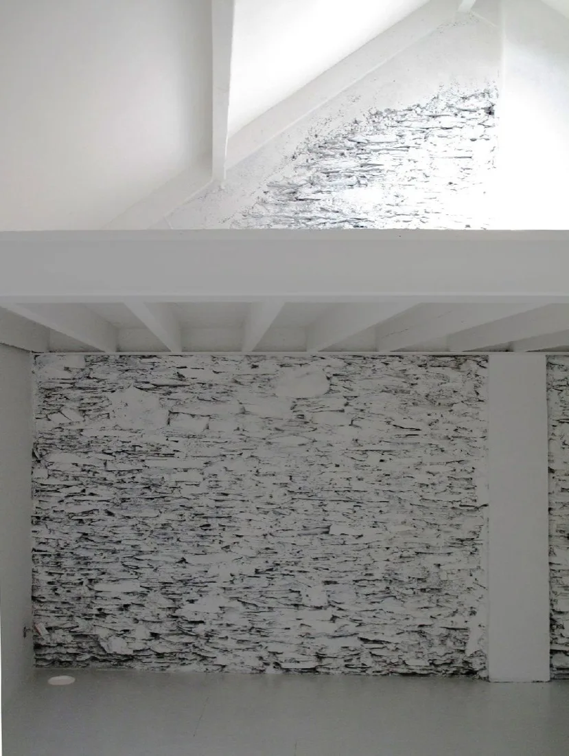 A minimalist white interior features a heavily textured painted stone wall beneath white structural beams and a vaulted ceiling.