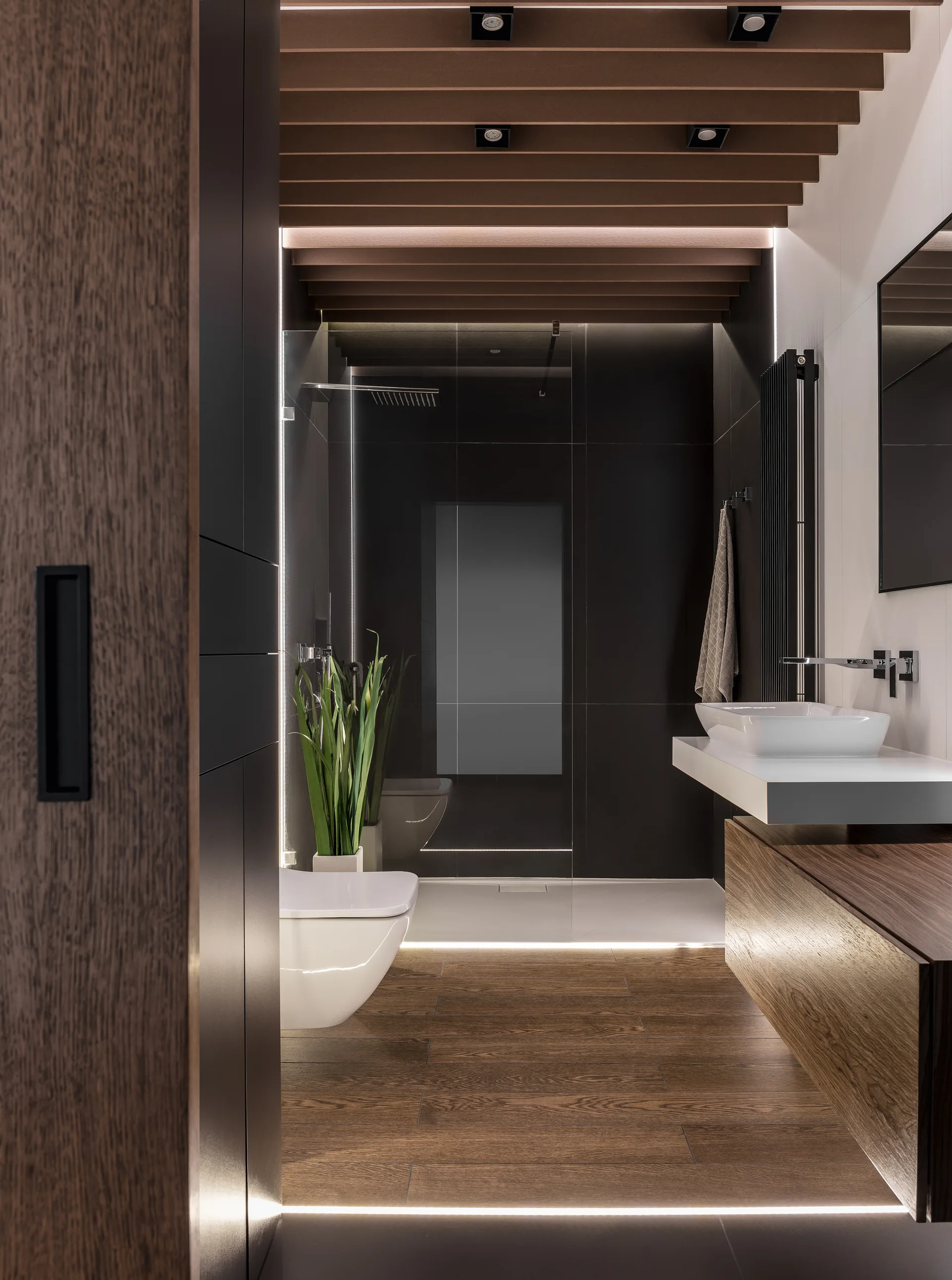 A modern bathroom features a dark tile walk-in shower, a wooden slat ceiling, a floating vanity, and floor-level linear LED lighting.