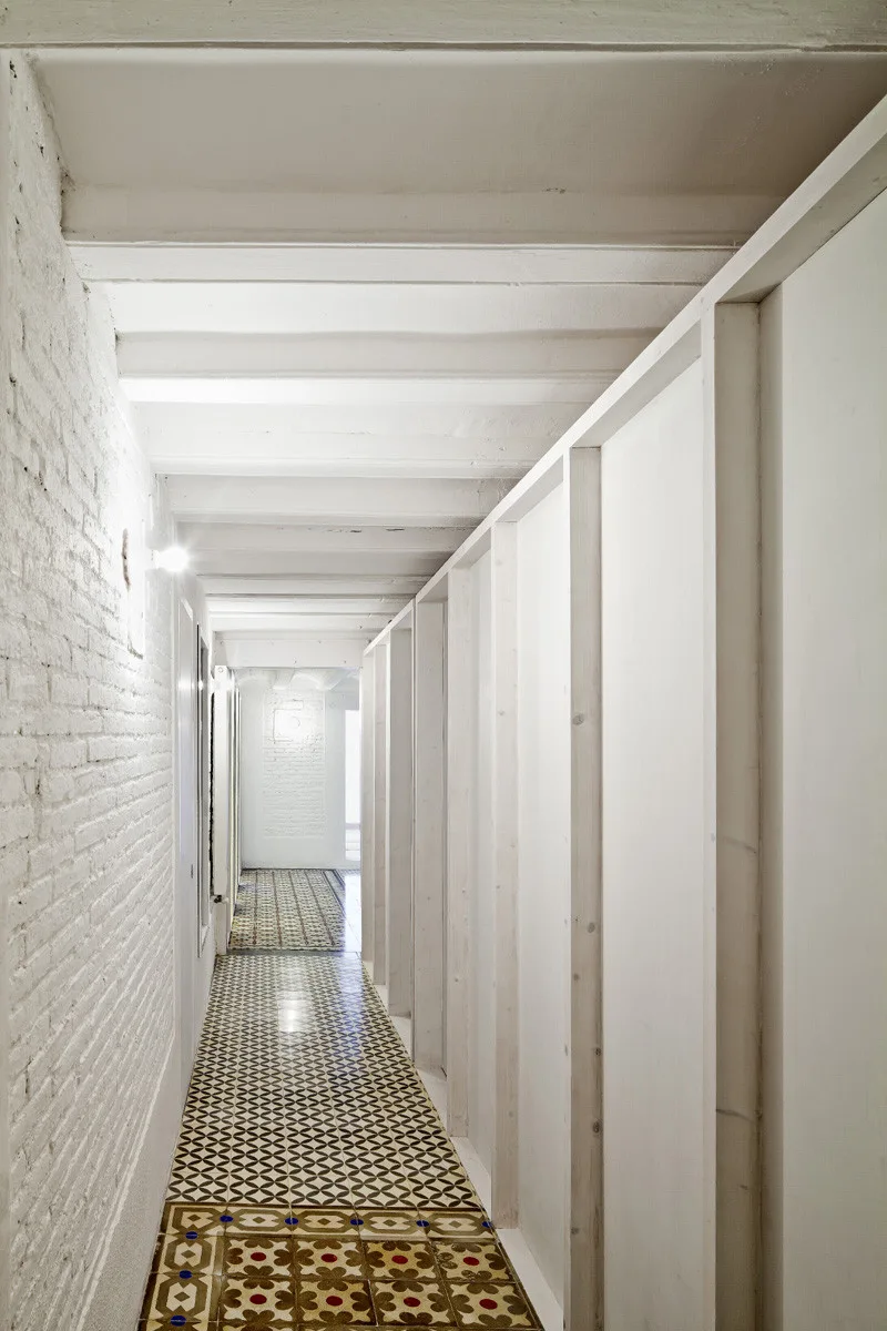A long white hallway featuring painted exposed brick walls, ceiling beams, and decorative geometric floor tiles.