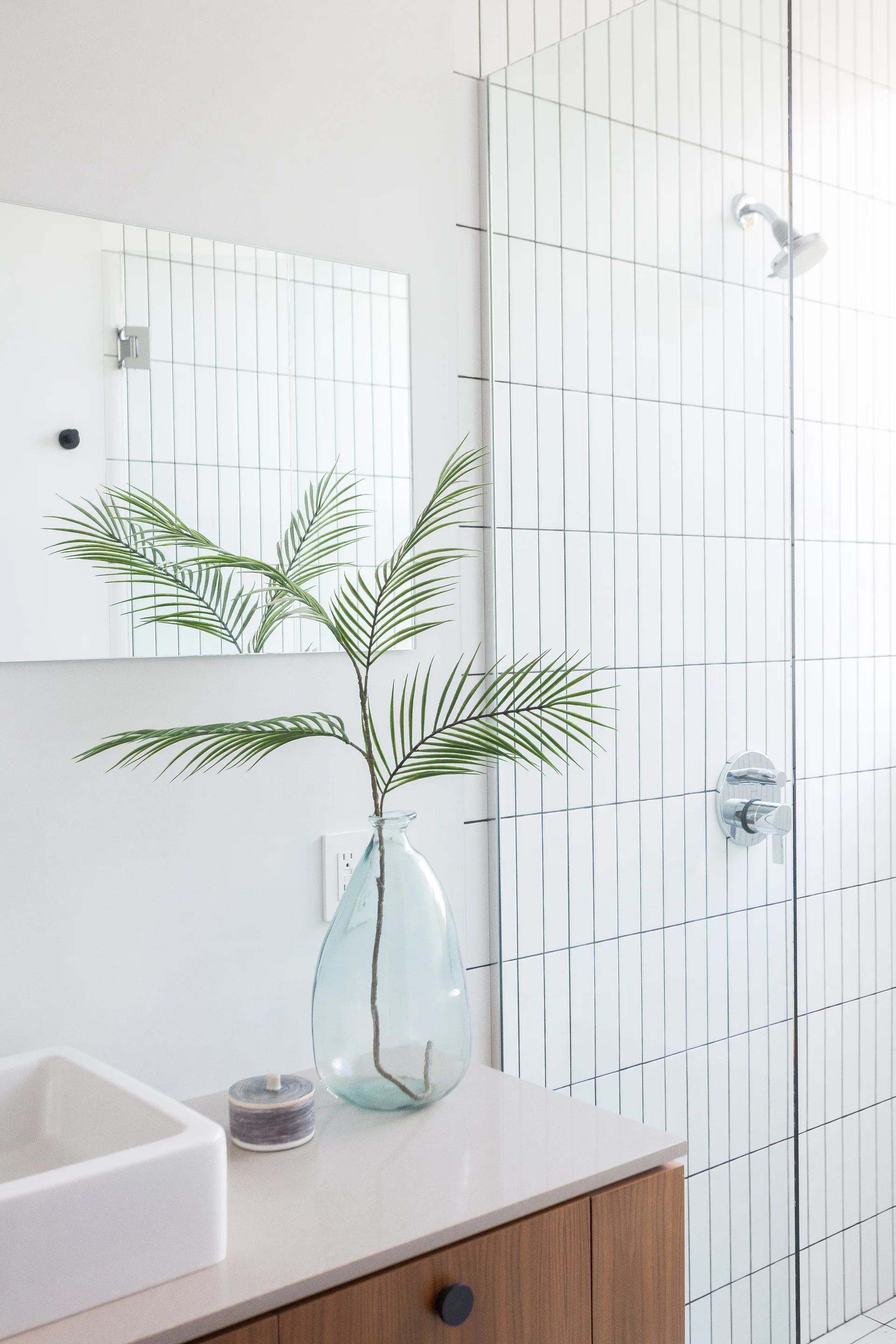 A modern wood vanity with a white vessel sink sits next to a glass shower featuring vertical white subway tiles and a chrome shower head.