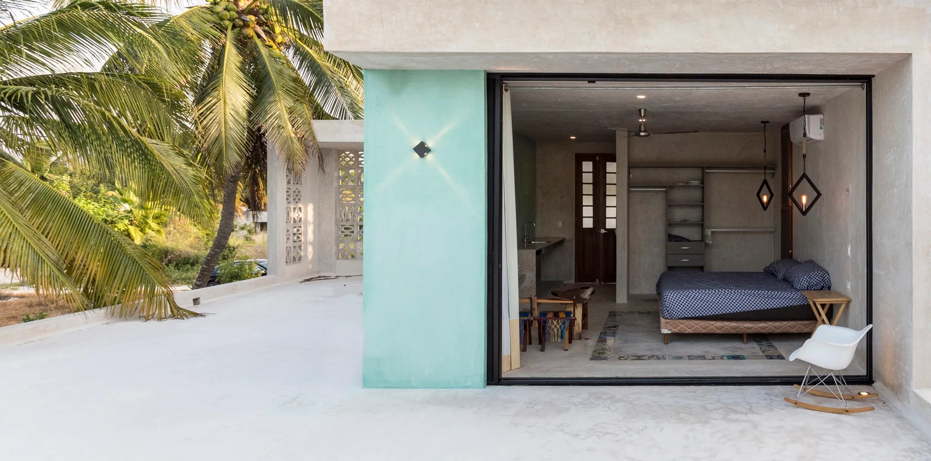 A concrete bedroom with a patterned bed opens directly onto an outdoor patio featuring a white rocking chair and a large palm tree.