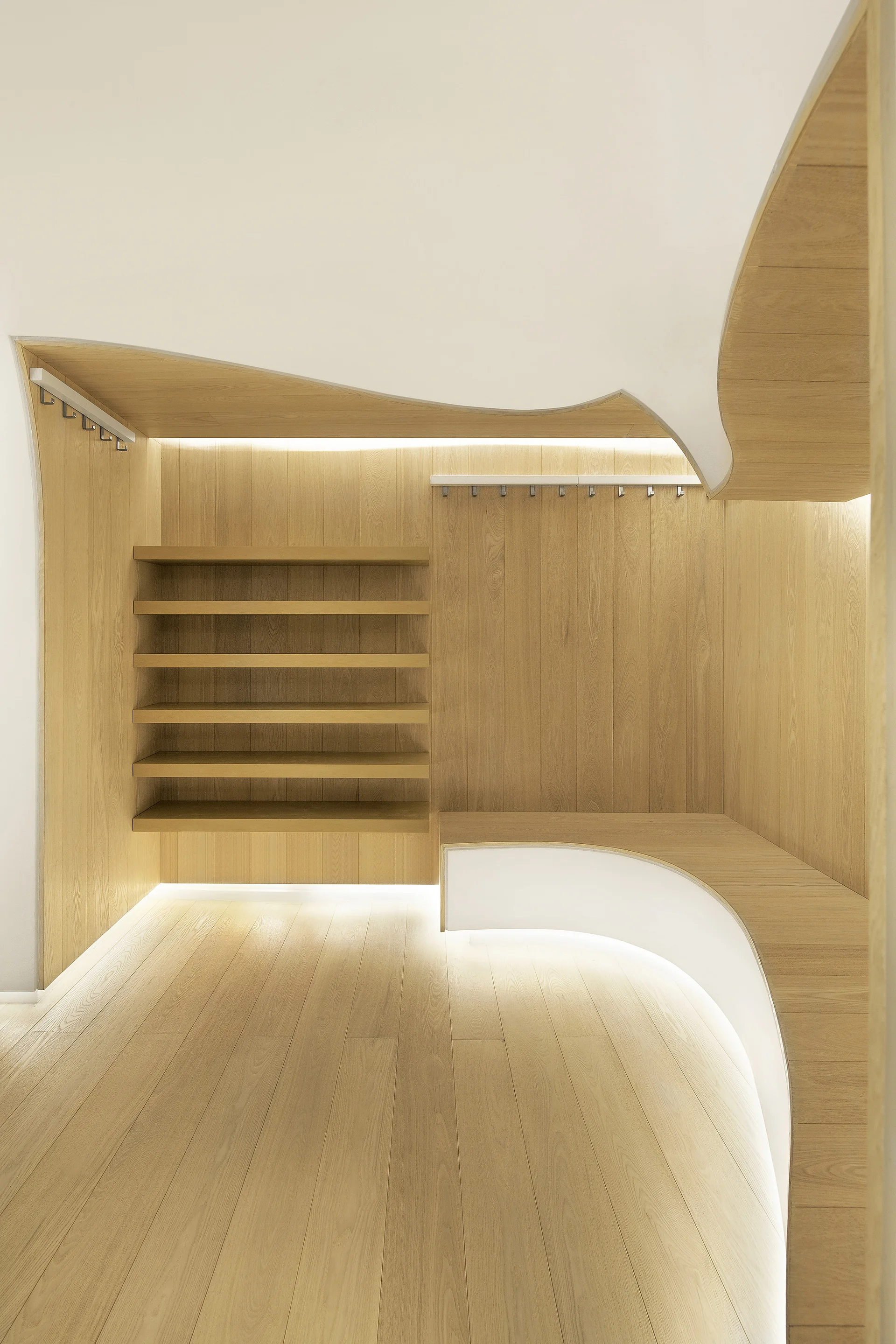 A minimalist light wood mudroom features custom built-in shelving, coat hooks, and a curved wooden bench illuminated by under-lighting.